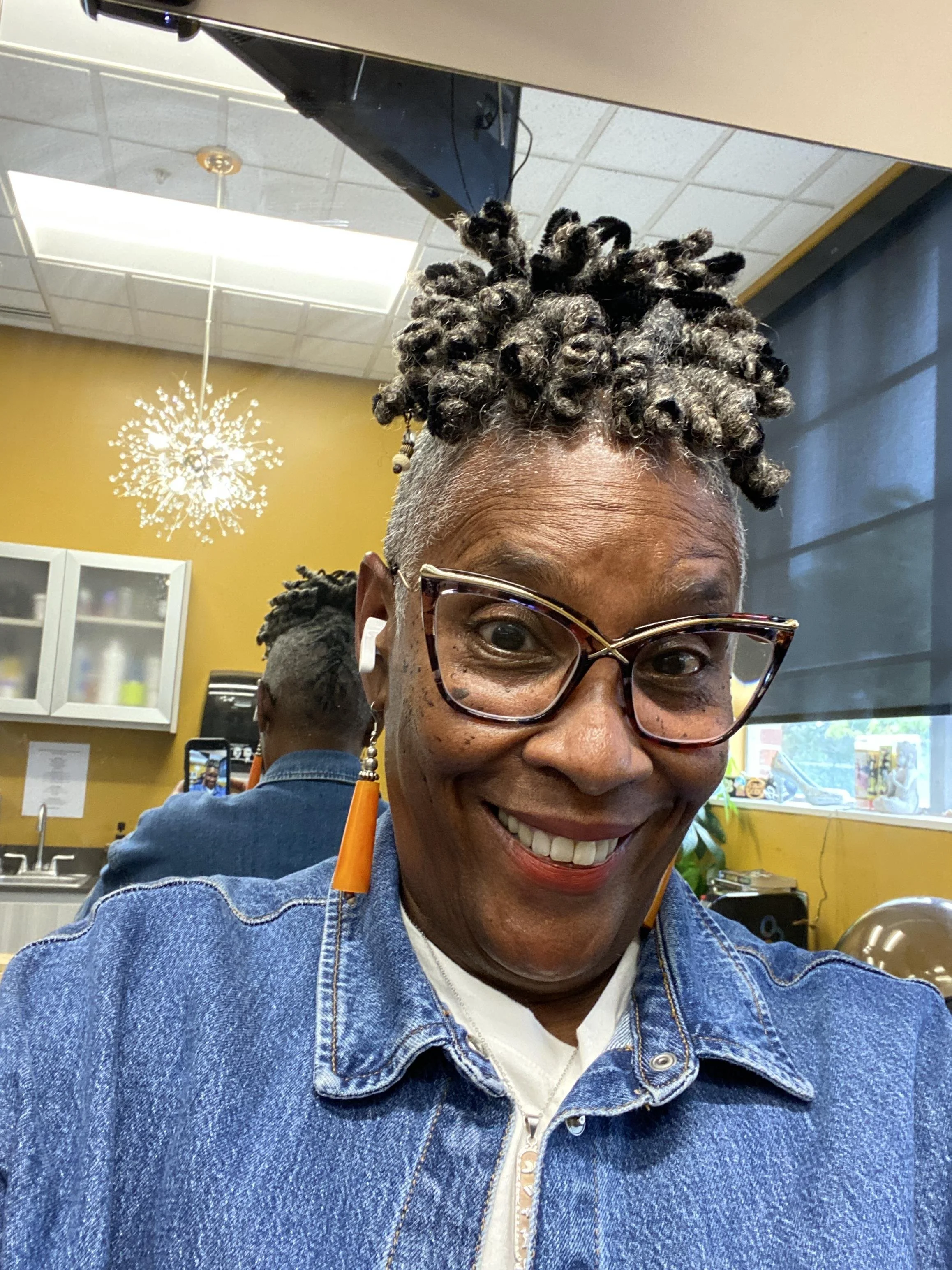 Smiling person with glasses and earphones wearing a denim jacket, standing indoors with a modern hairstyle, reflected in a mirror.