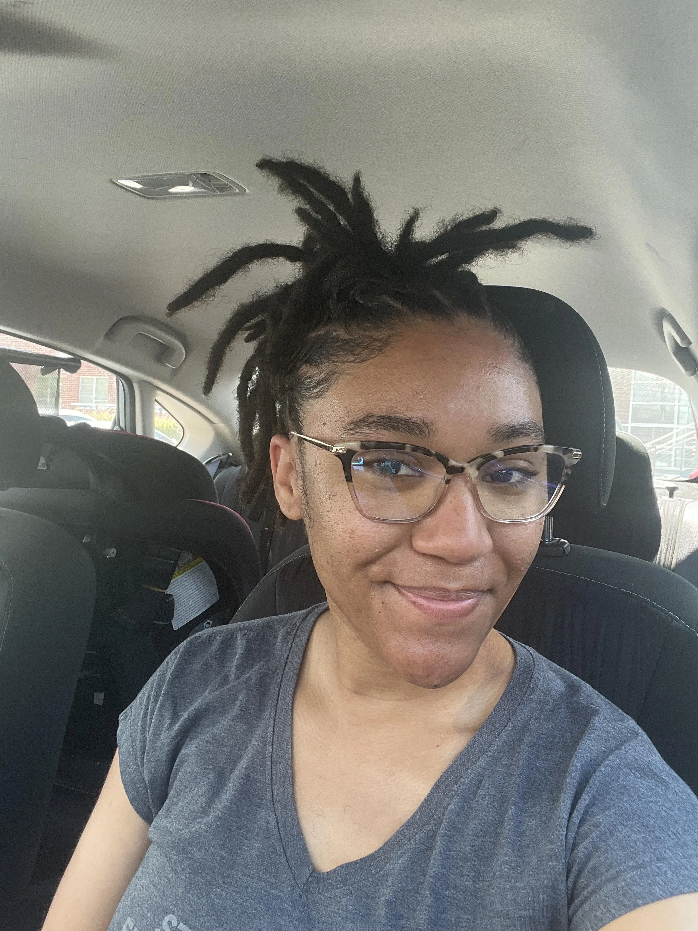 Person with glasses and dreadlocks sitting in a car, smiling.