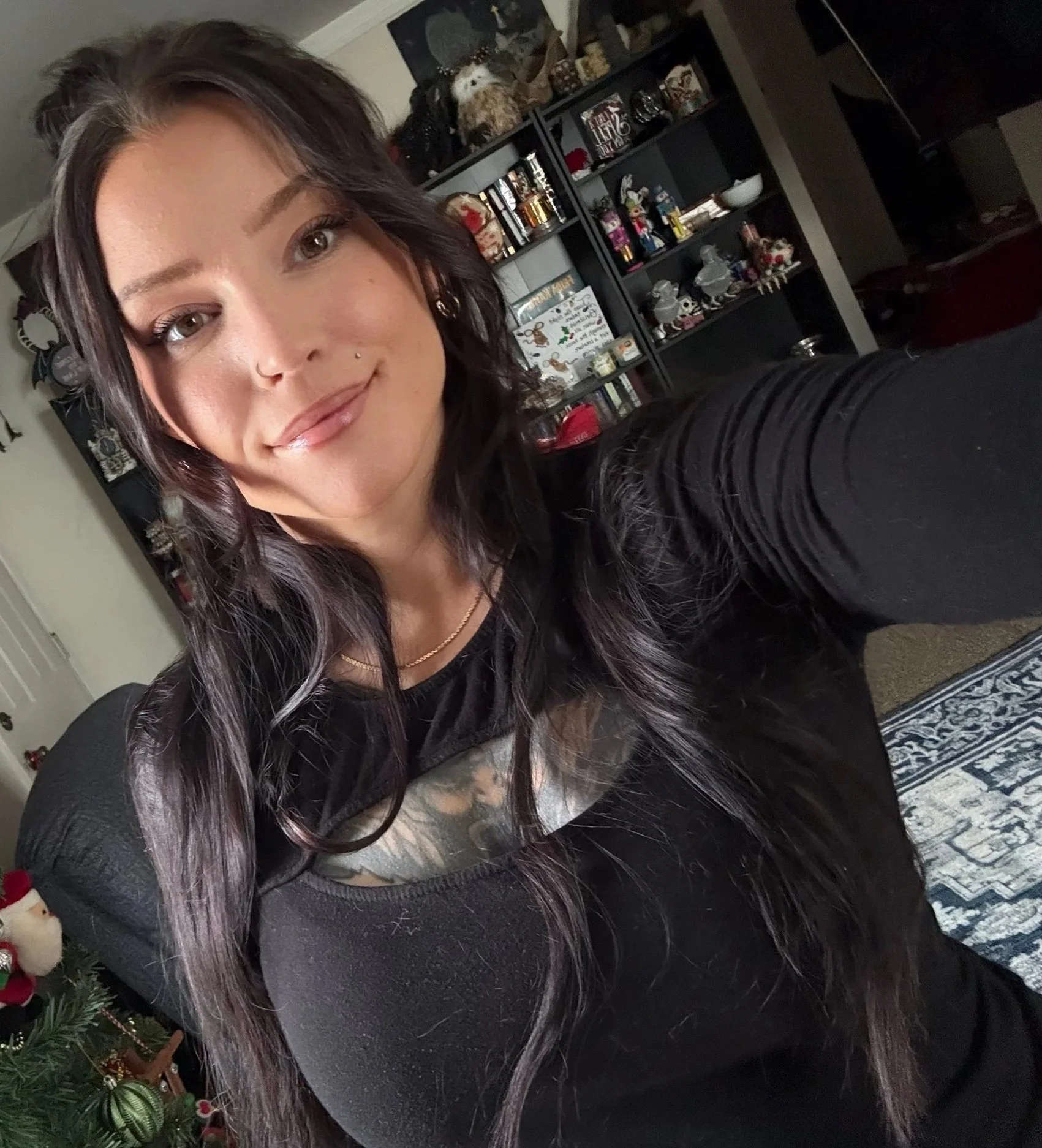 A woman taking a selfie indoors, with a black bookshelf filled with decorative items and books in the background, and a Christmas tree partially visible on the left.