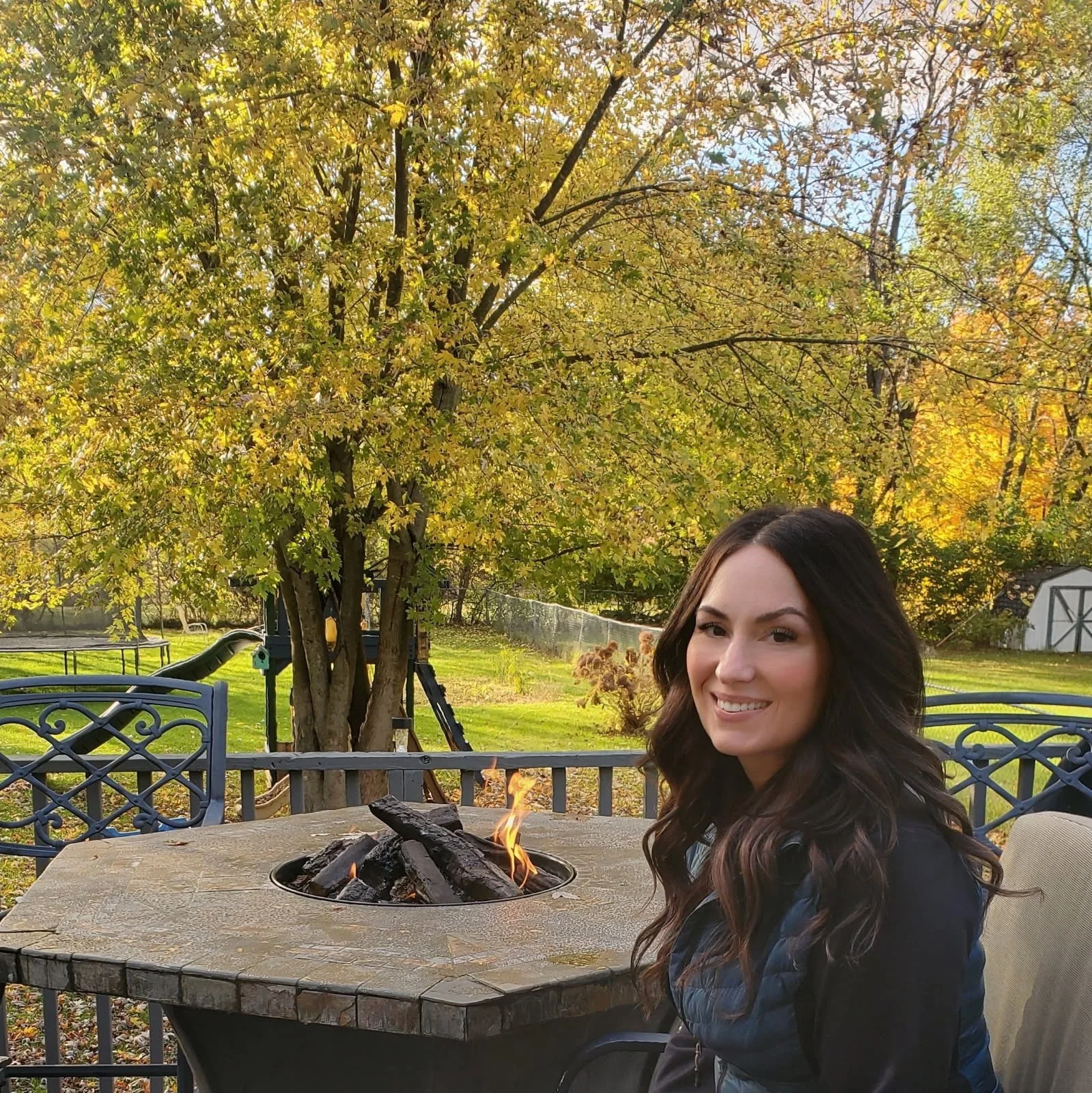 A woman with long dark wavy hair smiling while sitting at a patio table with a fire pit in front of her outdoors during autumn. Fall trees with yellow leaves are in the background.
