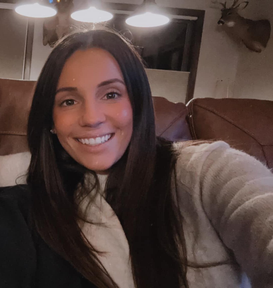 A woman with long dark hair smiling and taking a selfie on a leather couch in a cozy room with warm lighting and a deer head mounted on the wall.