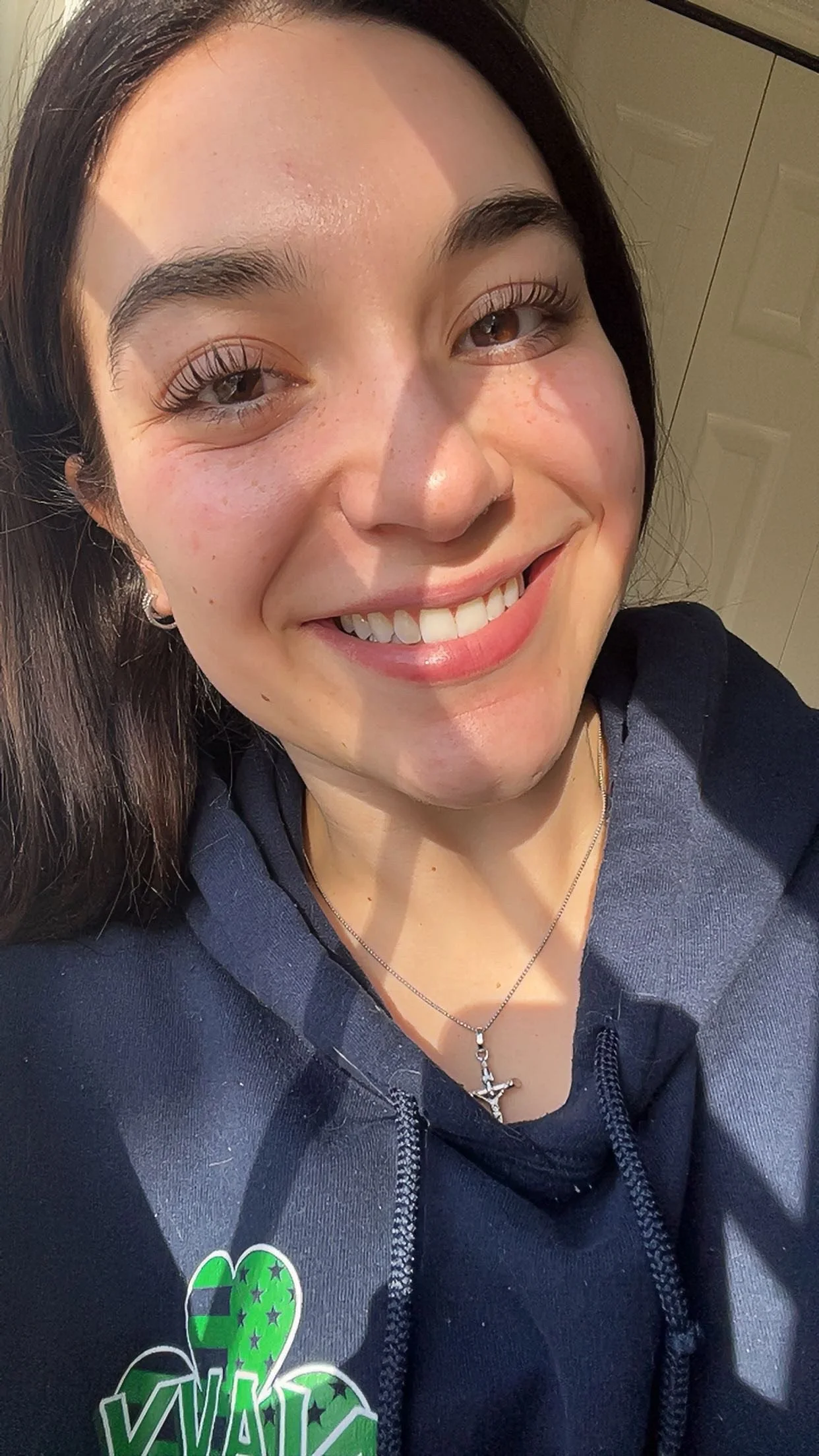 Close-up photo of a young woman with brown hair, smiling outdoors, wearing a dark hoodie with a green and black logo, a silver necklace with a cross pendant, and earrings, with sunlight on her face.
