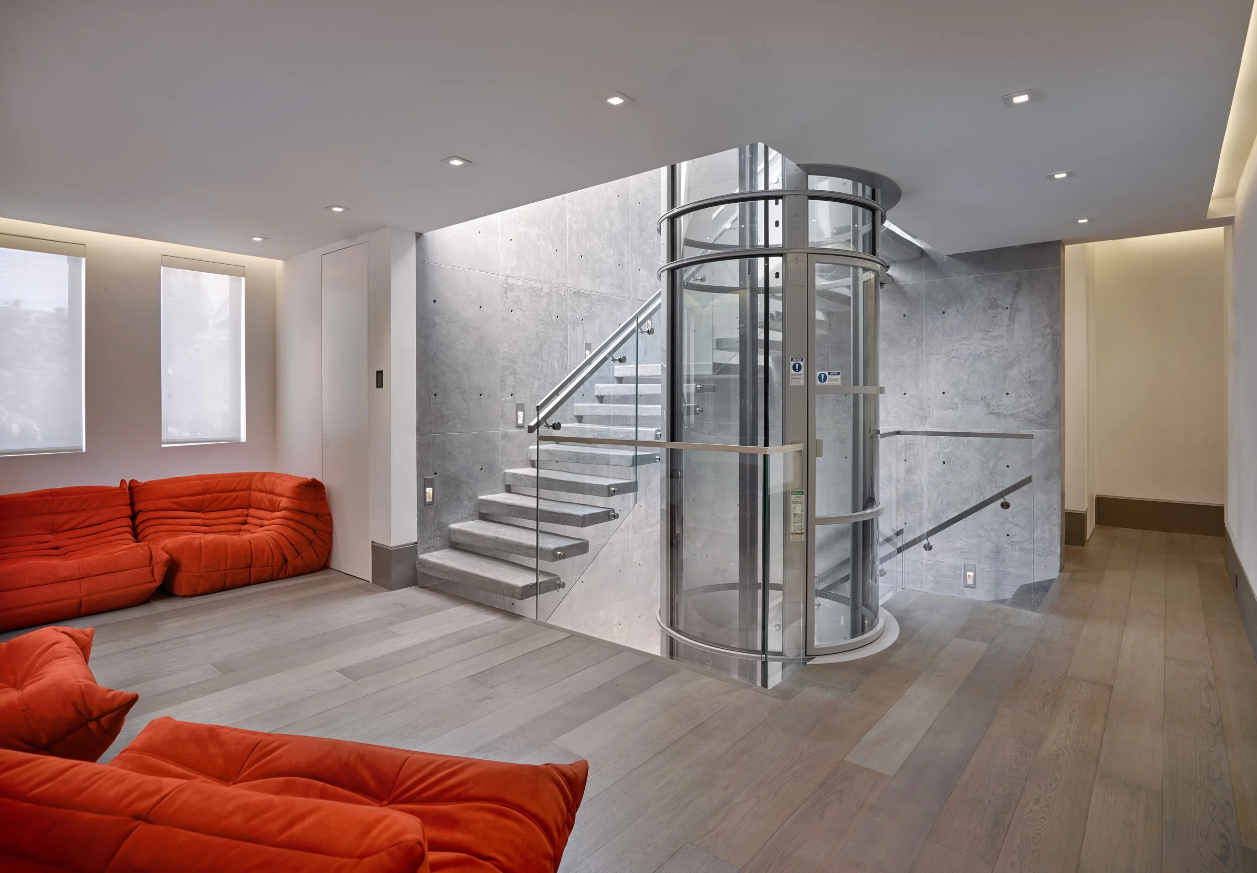 Modern interior with gray concrete walls, a glass elevator, wooden staircase, orange sectional sofas, and minimal lighting.