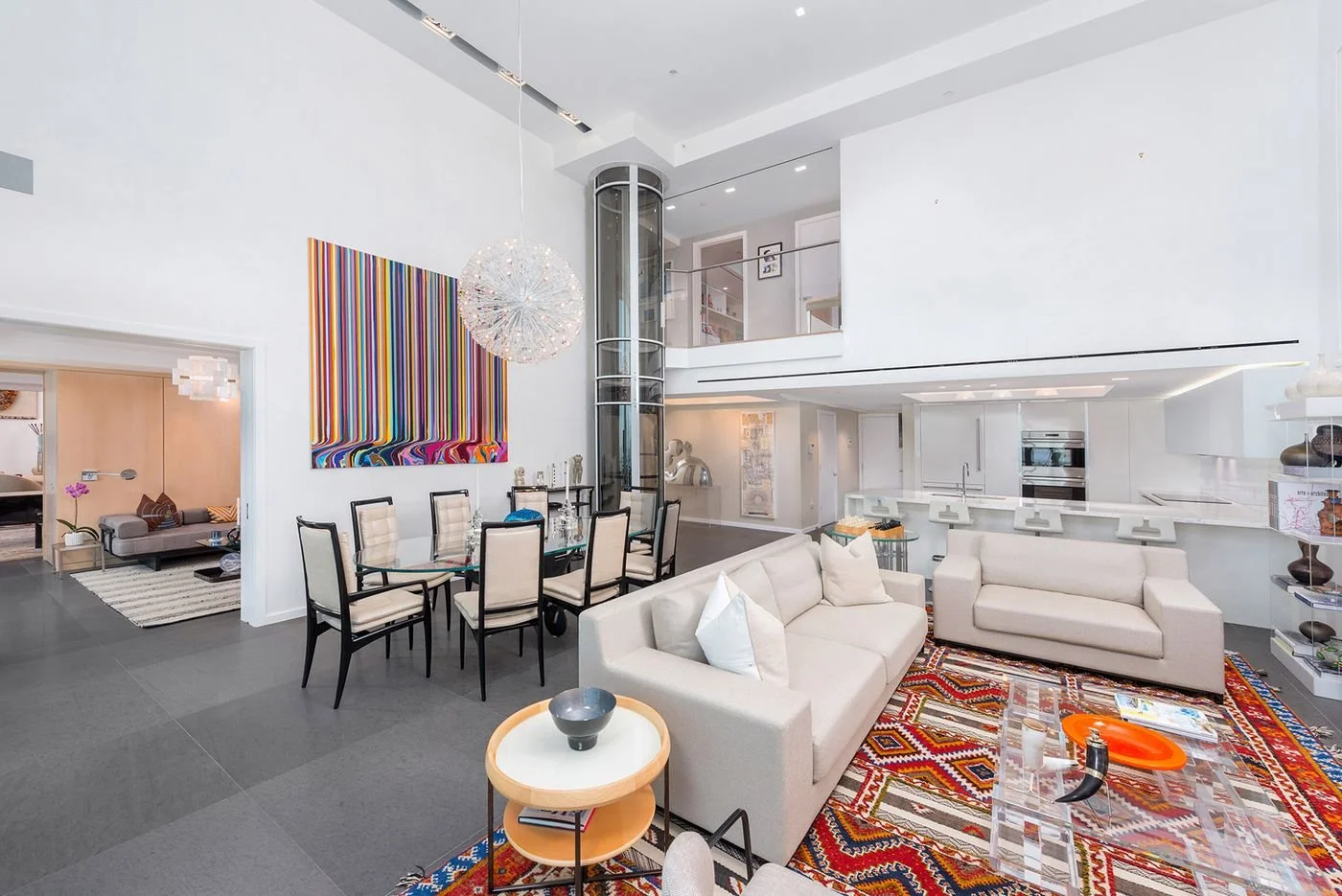 Modern open-concept living and dining area with white walls, a colorful striped wall art, a chandelier, a dining table with chairs, beige sofas, a glass coffee table, and contemporary kitchen in the background.