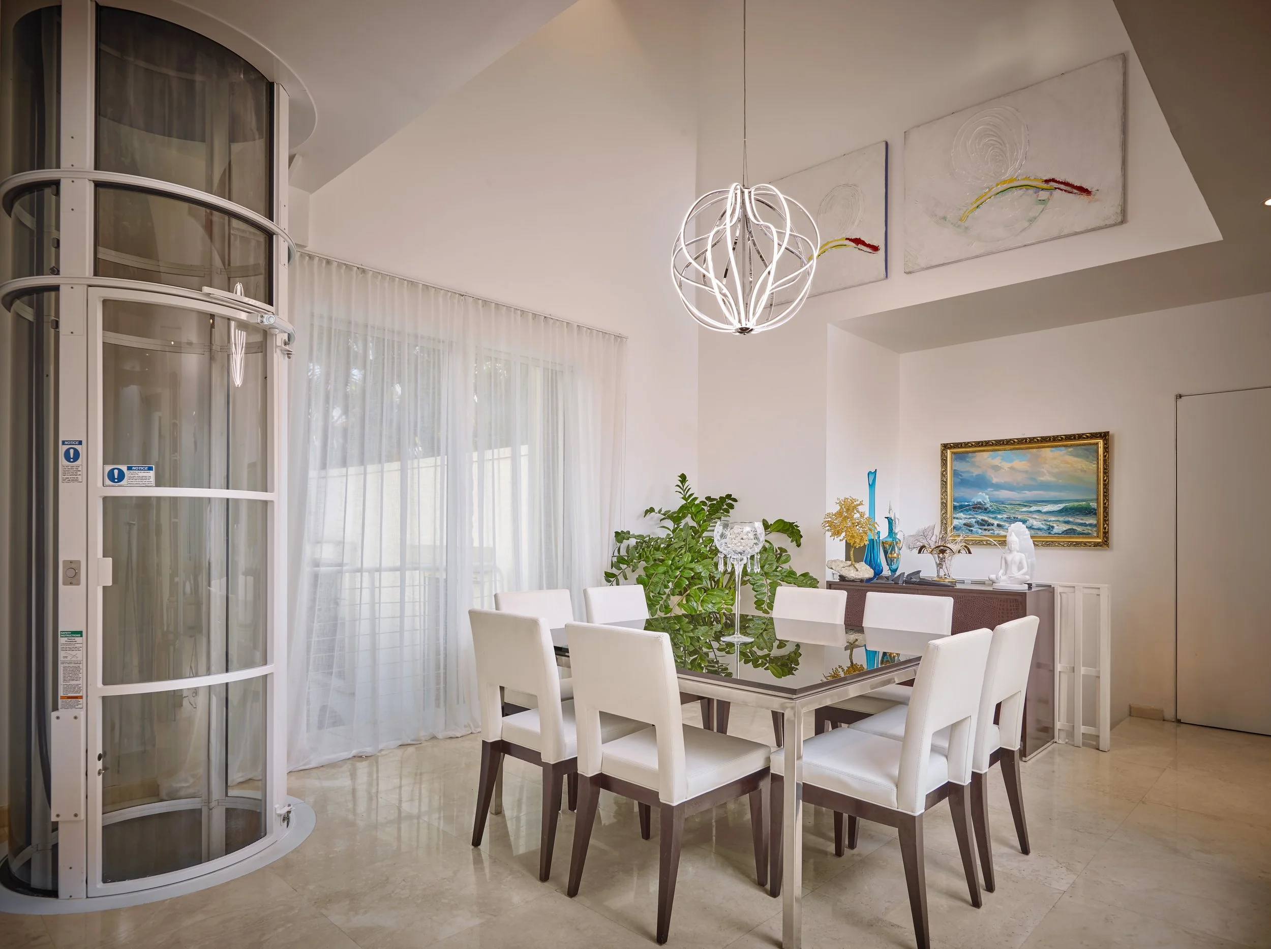 Modern dining room with white chairs around a glass-top table, a large window with sheer curtains, artwork on the walls, decorative vases, and a black cabinet.