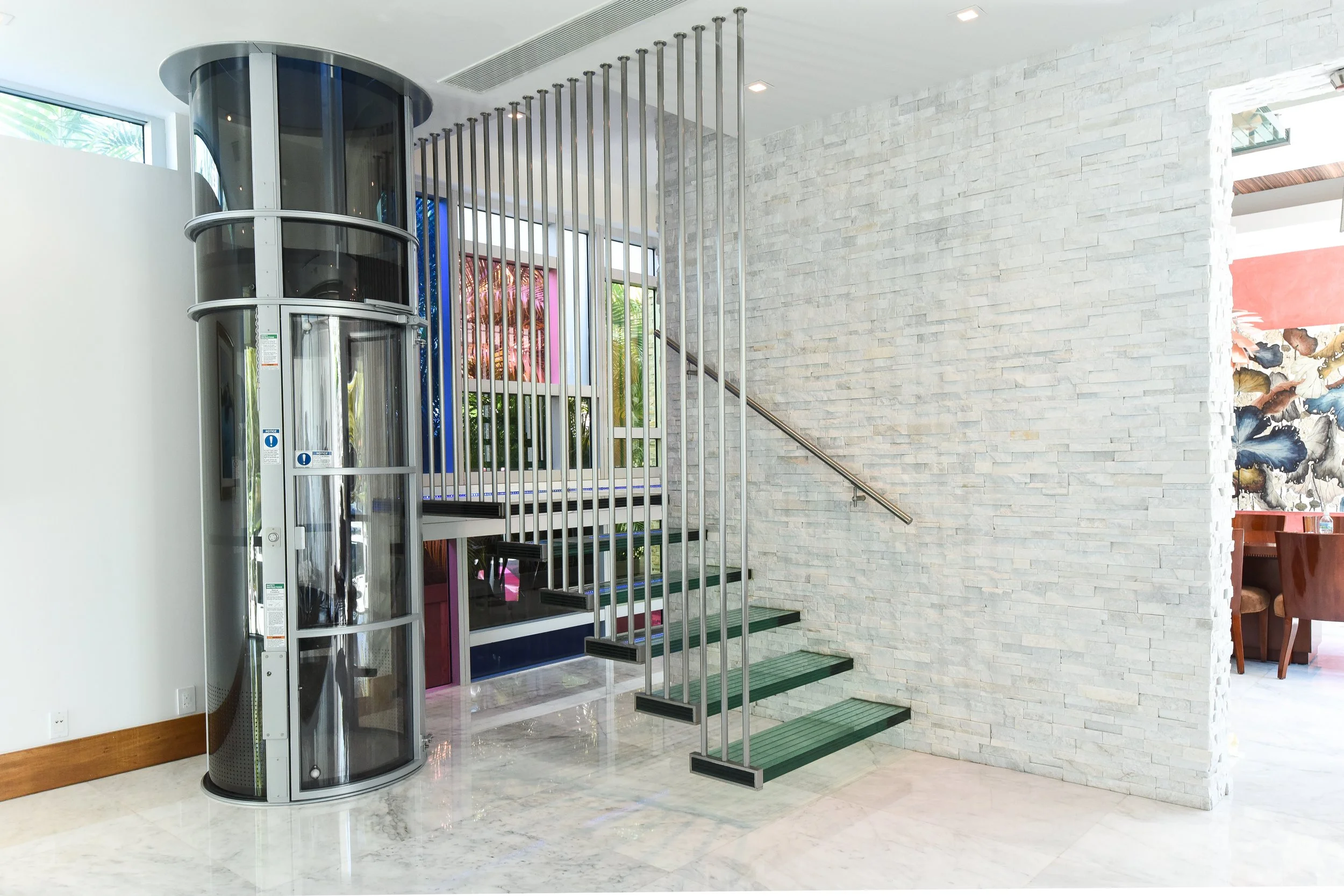 Indoor space with a glass elevator and staircase, white brick wall, large window, and a dining area with chairs and artwork in the background.