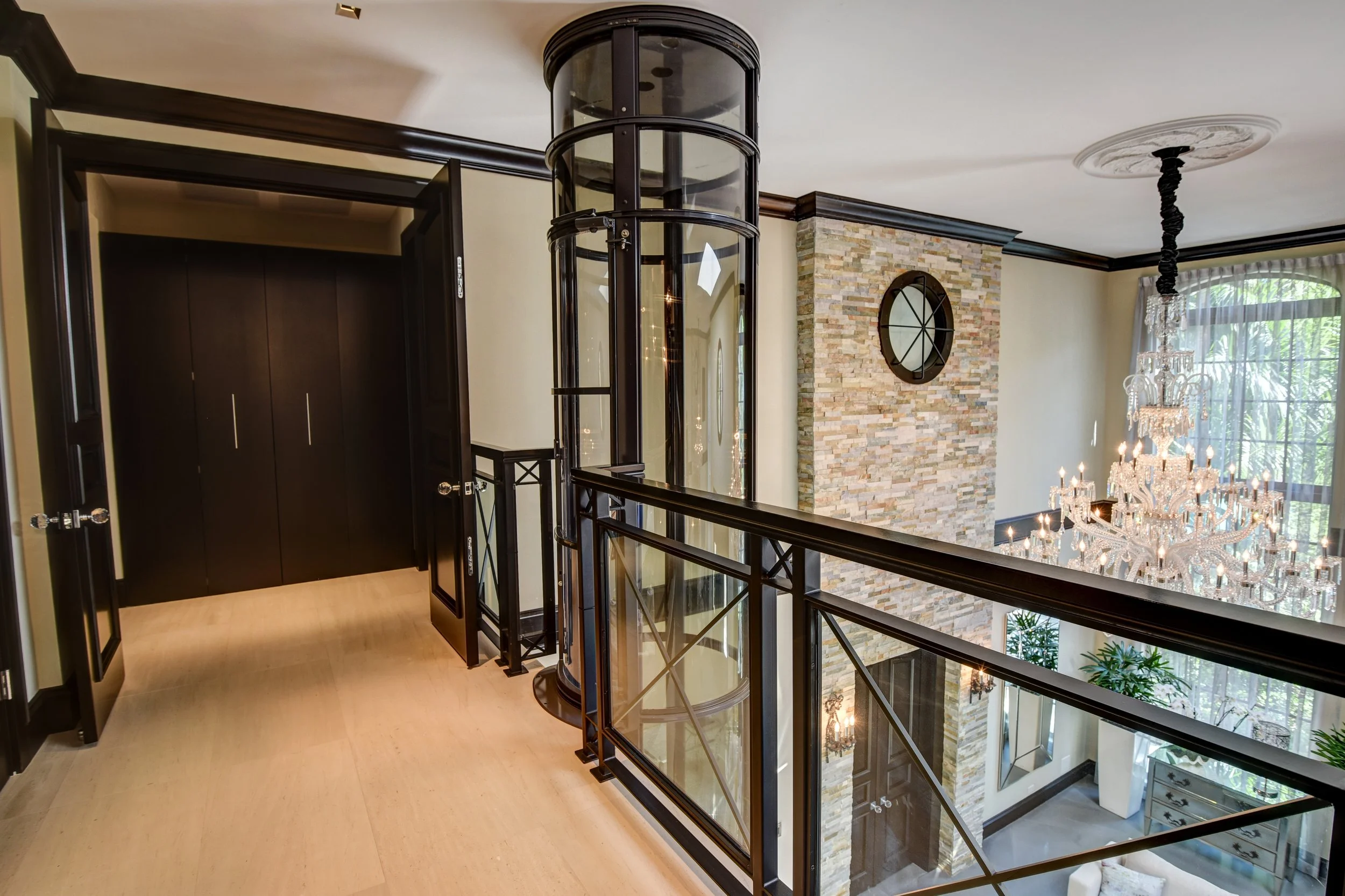 Interior view of a luxurious home's upper floor, featuring a curved glass elevator, a stone accent wall with a round mirror, a black chandelier, and large windows with drapes.