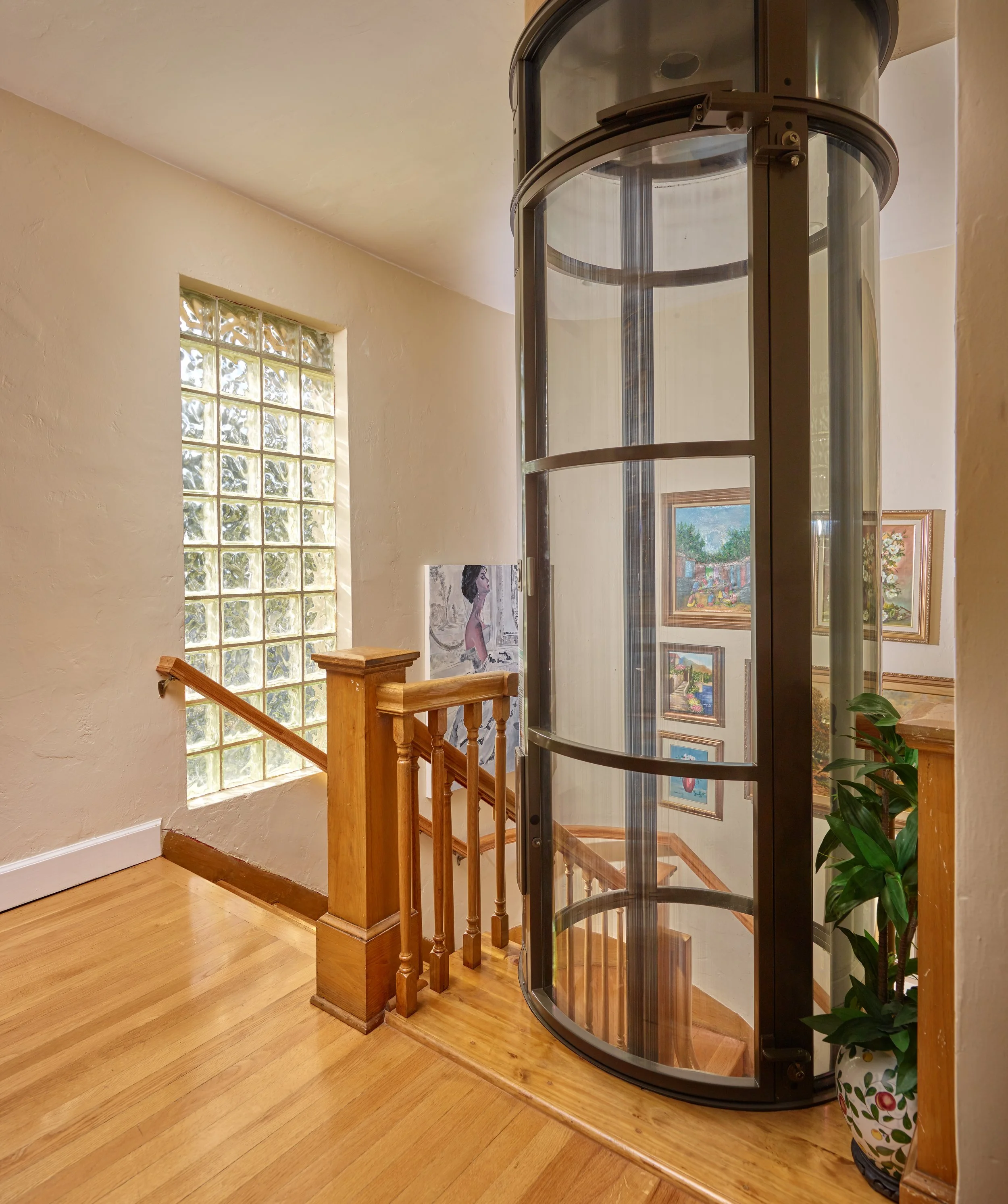 Interior view of a staircase in a house, showing a glass block window, a painting of a woman, framed artwork, a wooden staircase railing, a black spiral elevator or lift, and a potted plant with green leaves in a decorative vase.