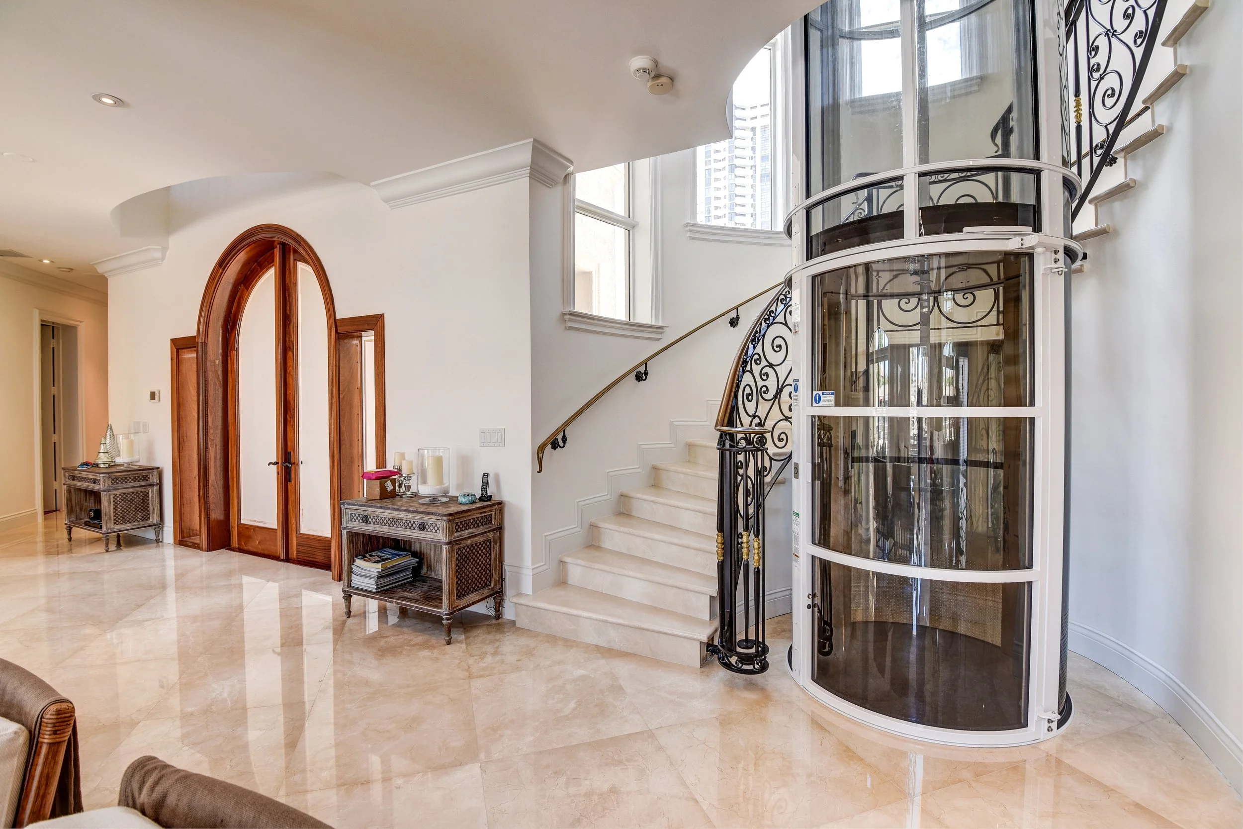 Interior of a luxurious home with cream marble floors, a curved staircase with ornate black wrought iron railing, a modern glass elevator with white framing, and a wooden door with an arched top.