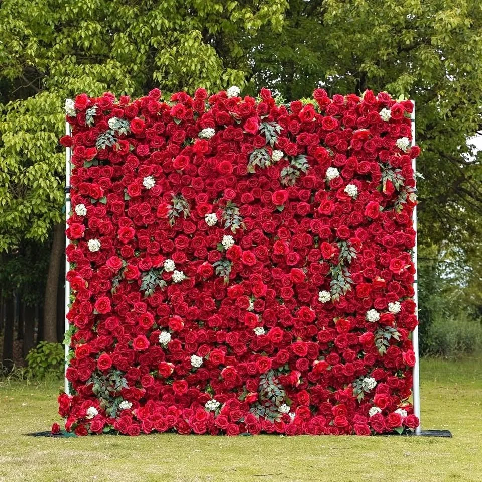 A large square floral display with numerous red roses, some white flowers, and dark green leaves, set outdoors on a grassy area with trees in the background.