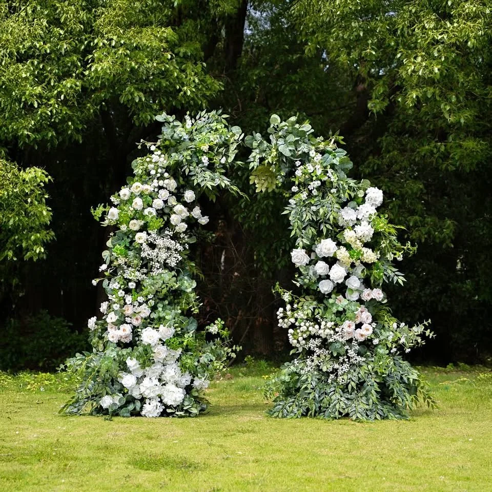 A floral arch made of white flowers and green foliage set outdoors on a grassy lawn with trees in the background.
