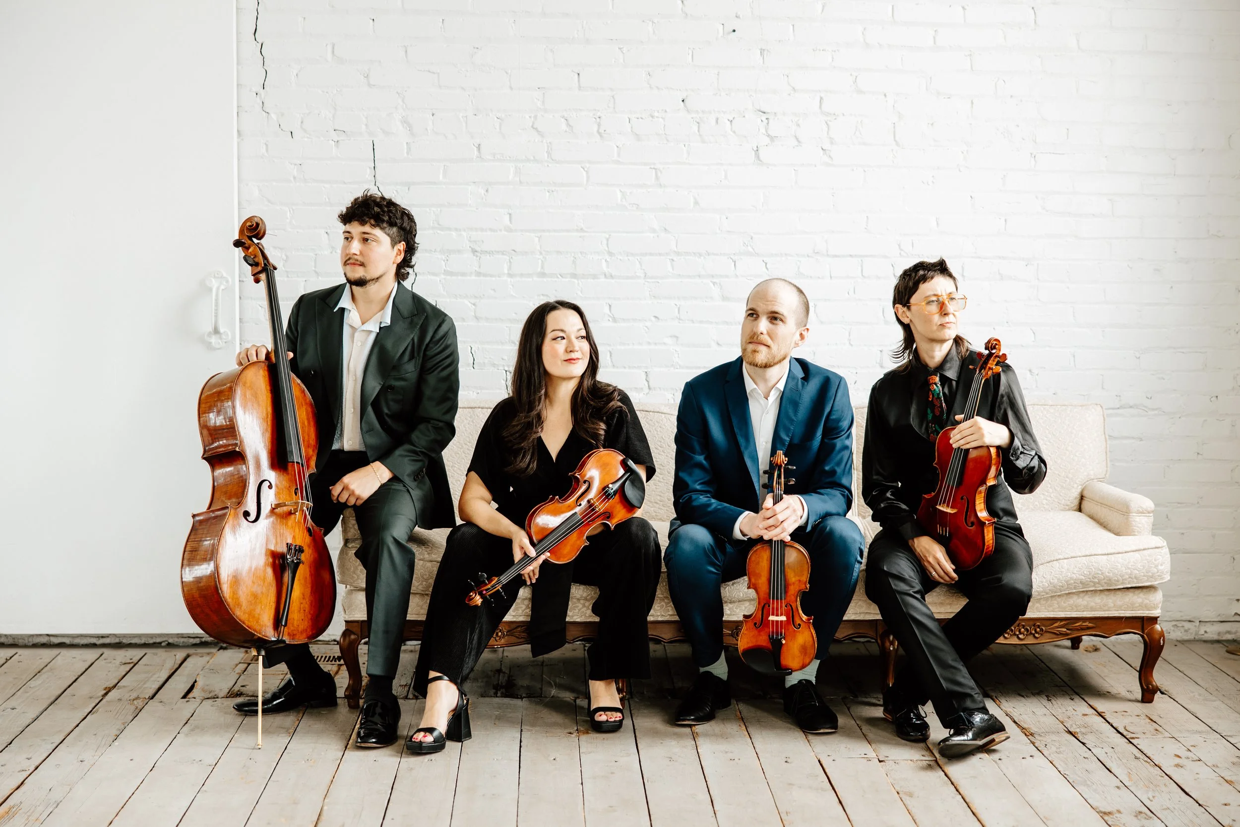 Thalea String Quartet sitting on a vintage sofa with a white brick wall background. Each holds a string instrument, including a cello viola, and violins, dressed in formal dark and blue attire.