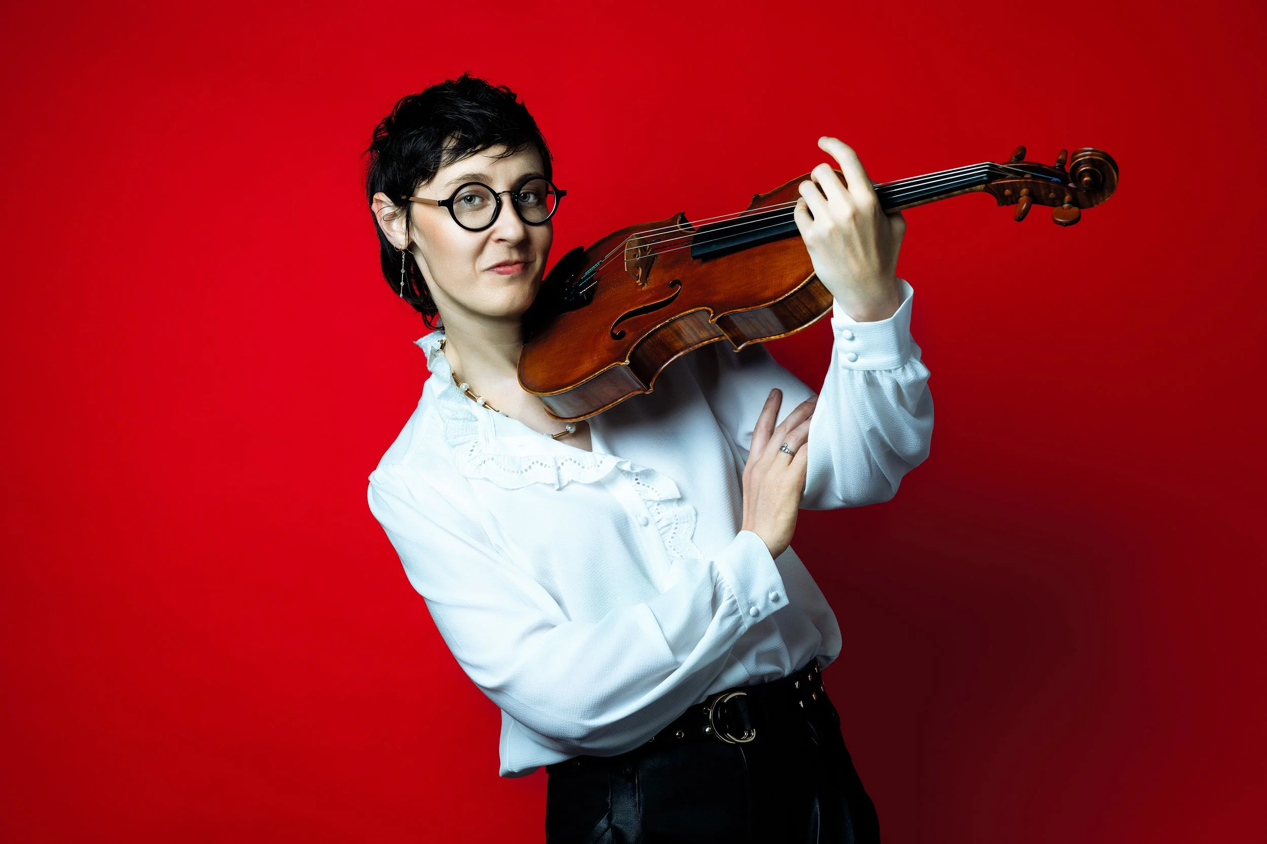 A woman with short black hair, glasses, and earrings holds a violin against a bright red background, looking confidently at the camera.