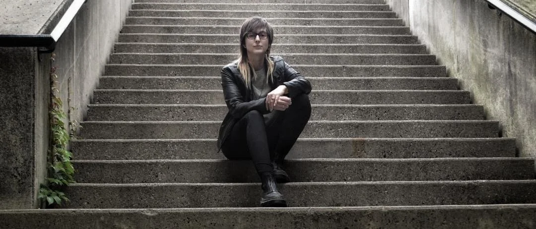 Rita Ushakova (Editor and Colourist based in Toronto) with glasses and long hair sitting on outdoor concrete stairs, wearing a black leather jacket, dark pants, and dark boots, looking cool for the camera.
