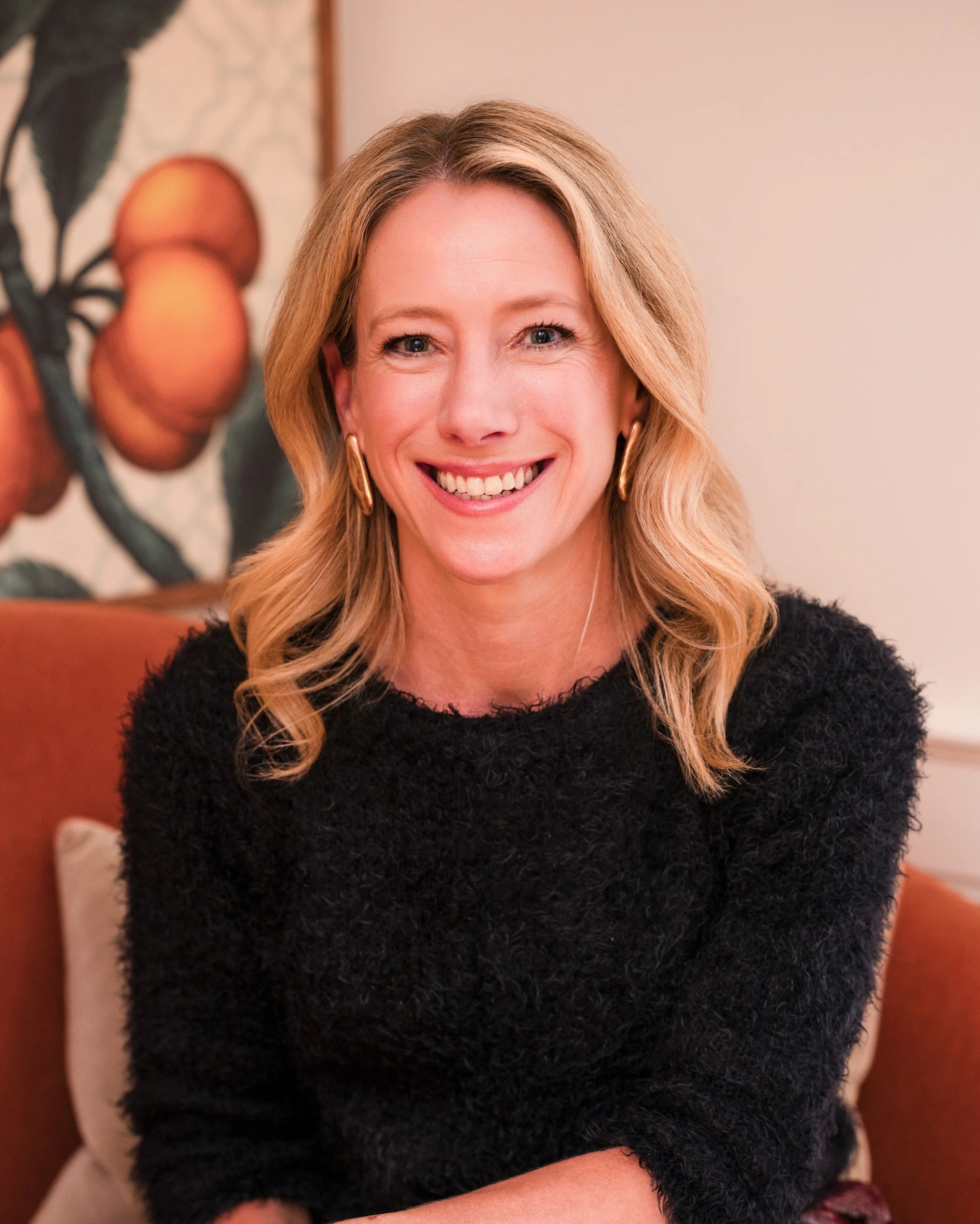 A woman sitting and smiling with blonde hair and wearing a black top.