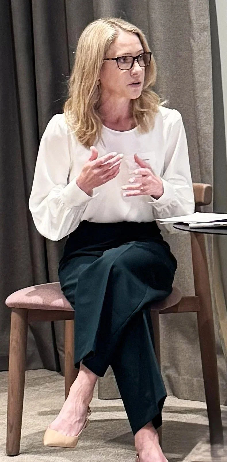 A woman with blonde hair and brown reading glasses wearing a white shirt and green trousers, sitting and speaking using hand gestures