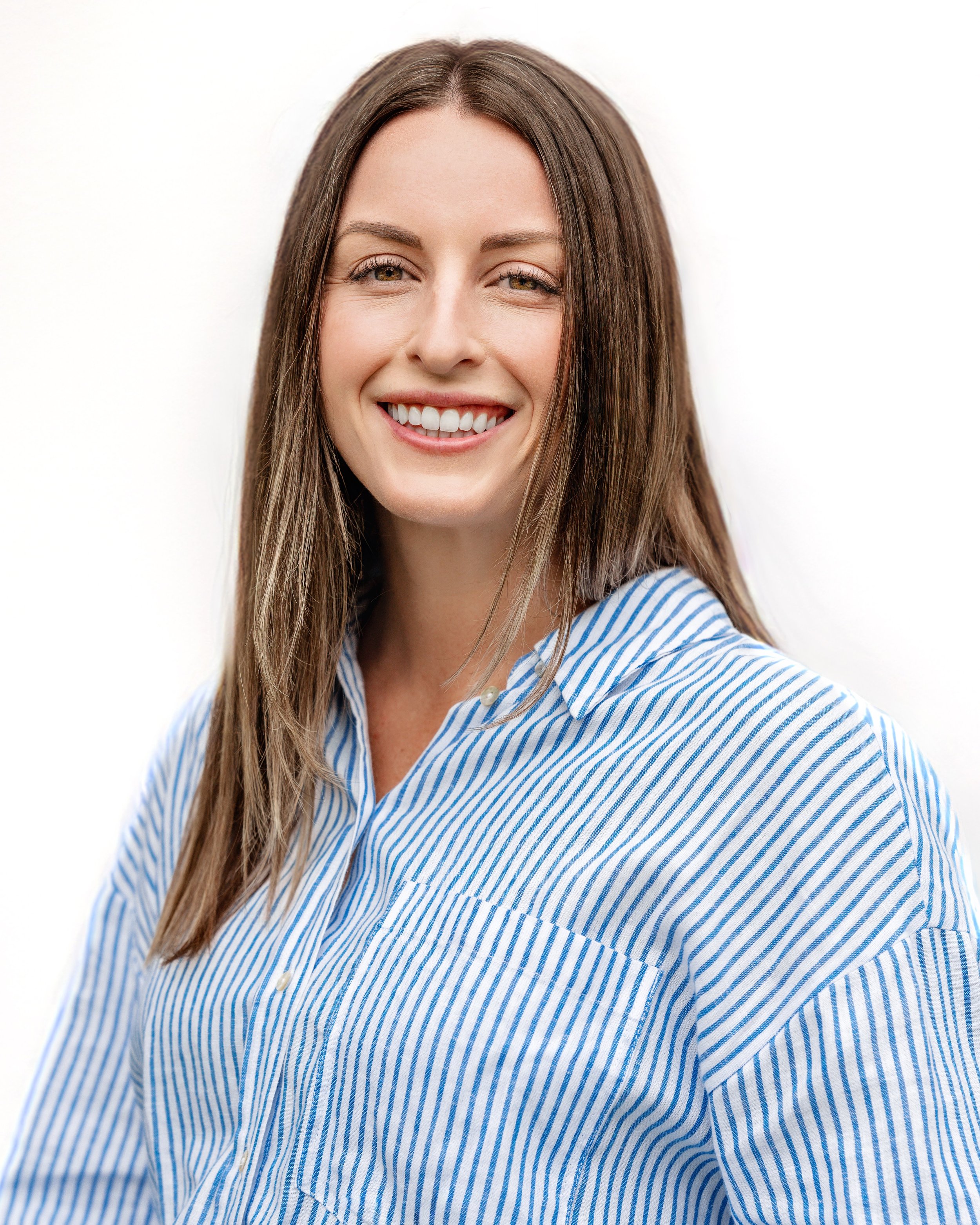 A woman with long brown hair, smiling, wearing a blue and white striped shirt, against a plain white background.