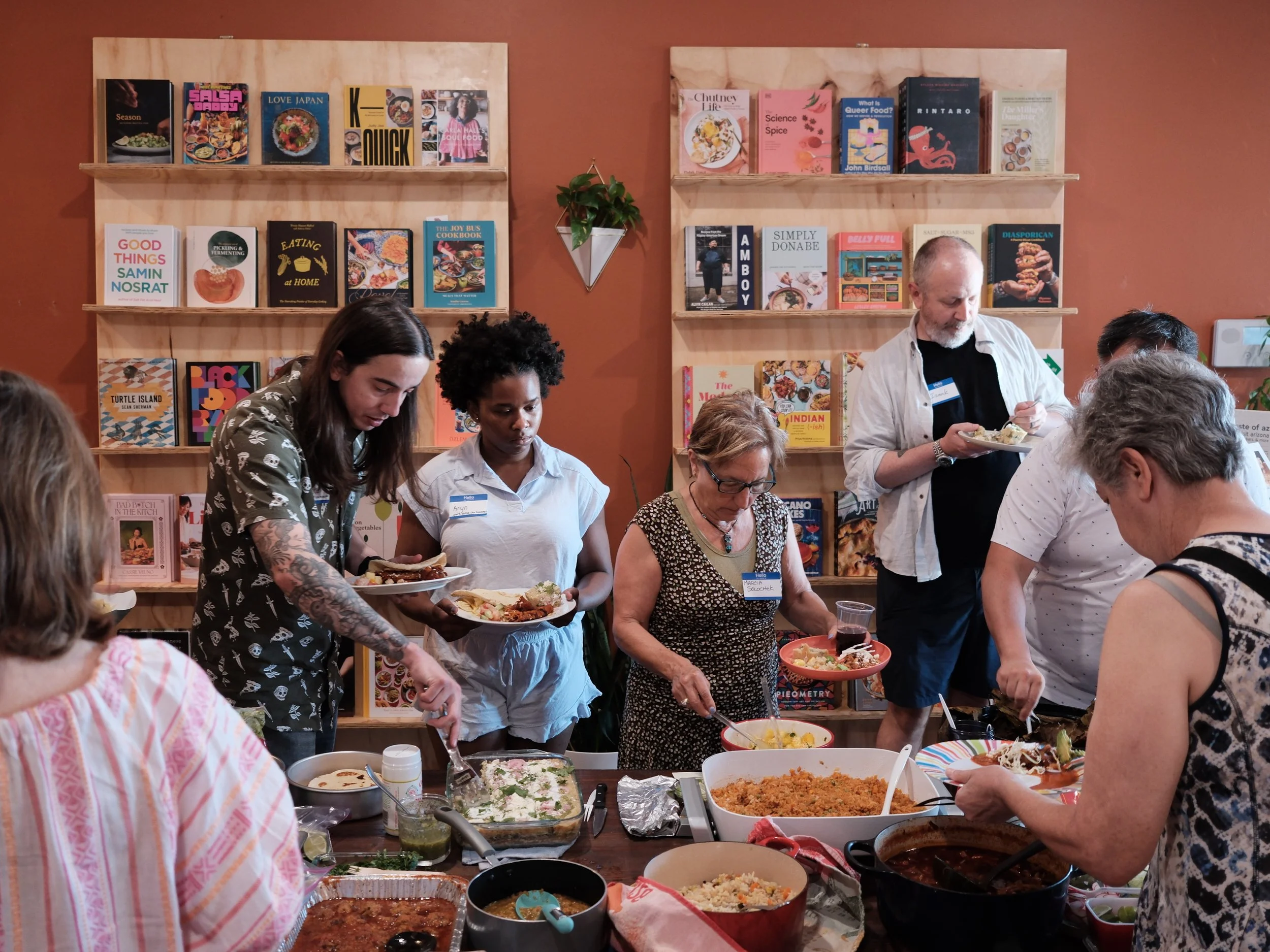 A group of people surround a table full of food for a cookbook club gathering at shea cheese lounge