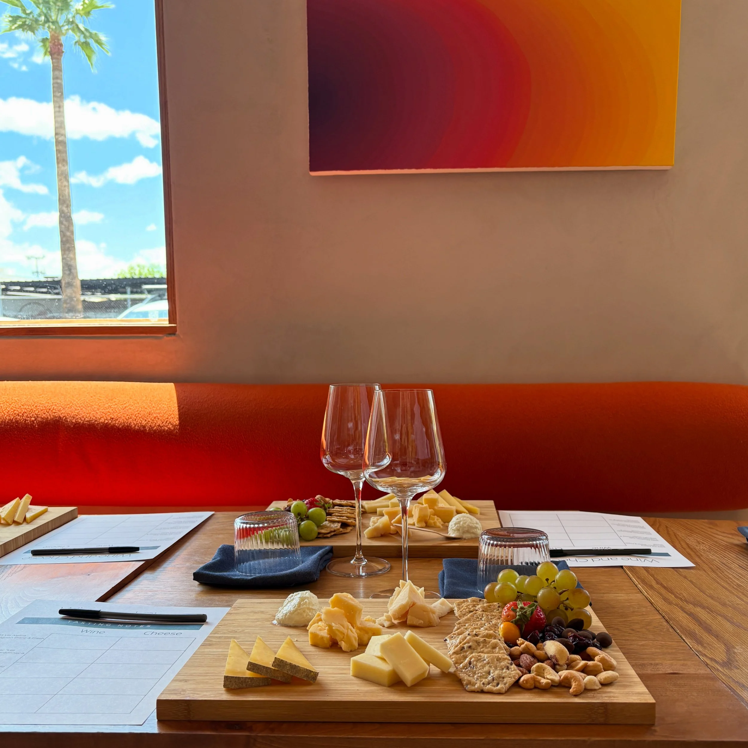 Cheese platter with various cheeses, grapes, crackers, nuts, strawberries, and chocolates on a wooden table with glasses and papers, in front of a window with a palm tree and blue sky.