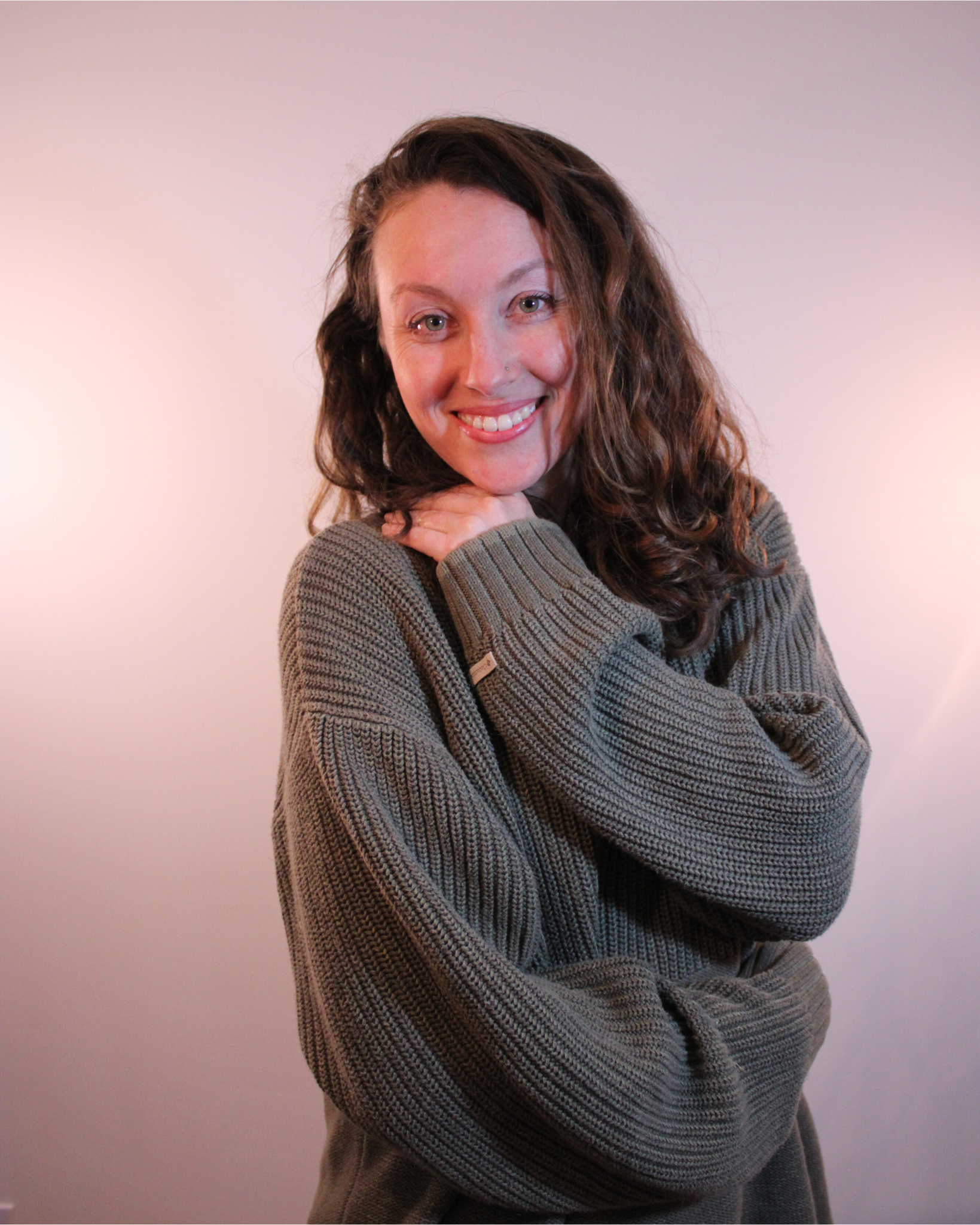 A woman with long, curly brown hair and light skin, smiling, resting her chin on her hands, wearing a gray knit sweater, against a plain light-colored background.