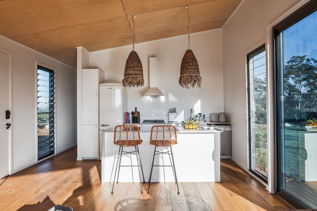 Sunlit modern kitchen with white cabinets, a central island with two wicker bar stools, wooden ceiling, and large windows showing outdoor greenery.