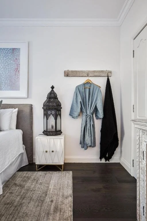 A bedroom corner with a bed partially visible on the left, a nightstand with a decorative lantern, a picture on the wall, and a robe hanging on a hook on the wall next to a black item, possibly a towel or piece of clothing.