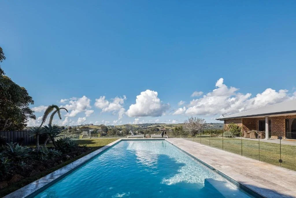 Long swimming pool with clear blue water, surrounded by a concrete patio, a grassy lawn, a house with a brick wall, and a scenic view of a cloudy sky and distant hills.