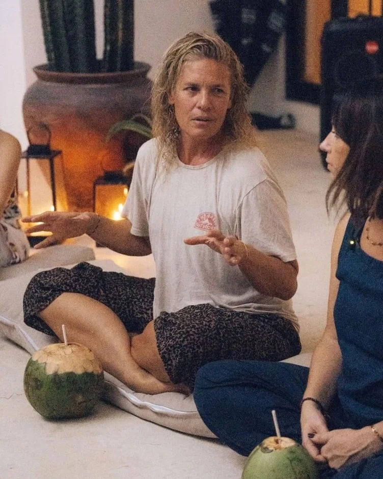 Two women sitting cross-legged on the floor with coconuts and drinks in front of them, engaged in conversation inside a cozy, dimly-lit room with large potted plant and warm lighting.