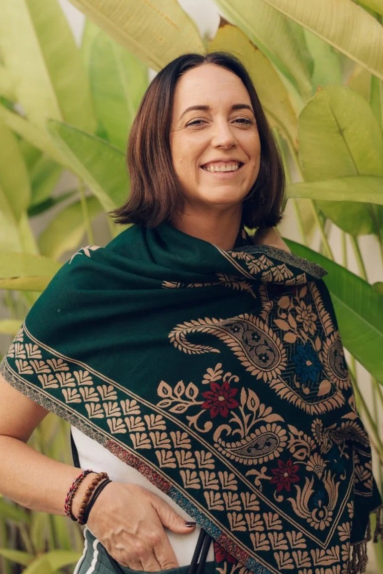 A woman smiling outdoors among large green banana leaves, wearing a dark green shawl with beige, red, and blue floral and paisley patterns.