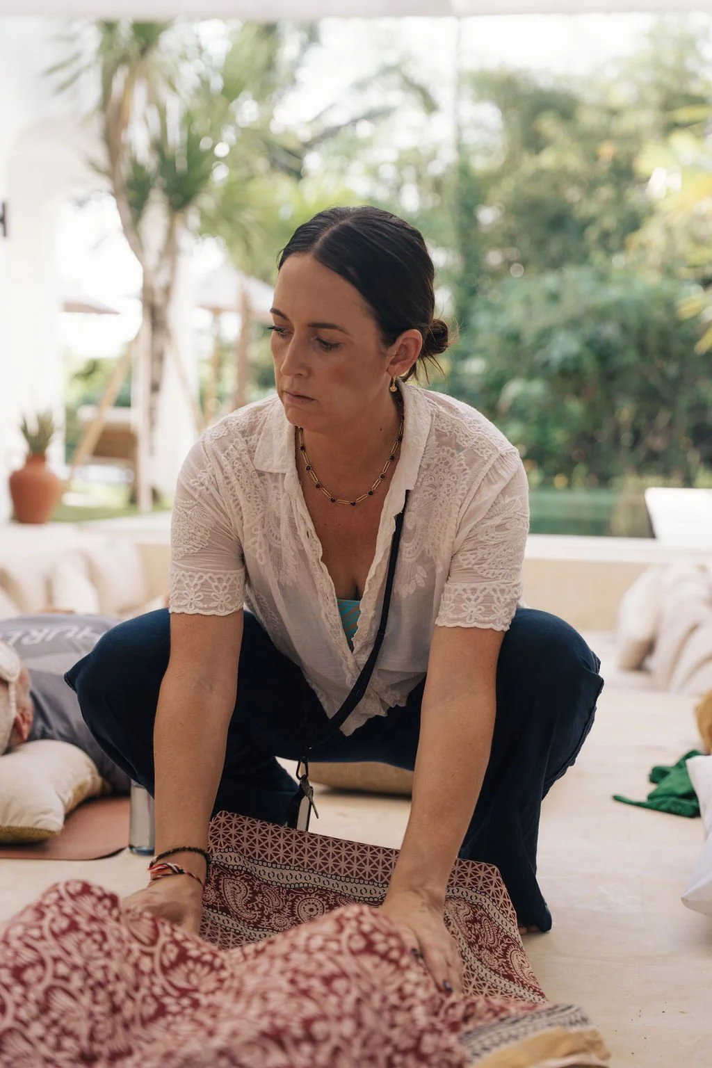 A woman with dark hair tied back, wearing a white lace blouse and a necklace, is squatting on a floor indoors. She appears to be tending to or interacting with a person lying down, surrounded by cushions and personal belongings. The background shows large windows with a view of trees and greenery outside.
