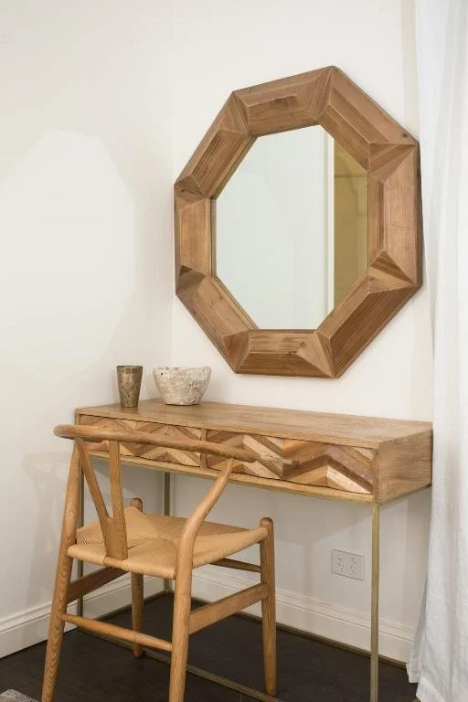 A wooden octagonal mirror hangs above a wooden desk with decorative vases, accompanied by a wooden chair in a corner with white walls and a curtain.