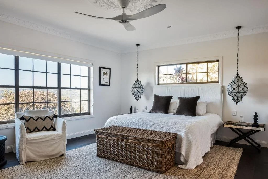 A bedroom with a large bed, dark pillows, a woven storage bench, a white armchair with a black and white pillow, hanging bedside lamps, large windows, and a ceiling fan.