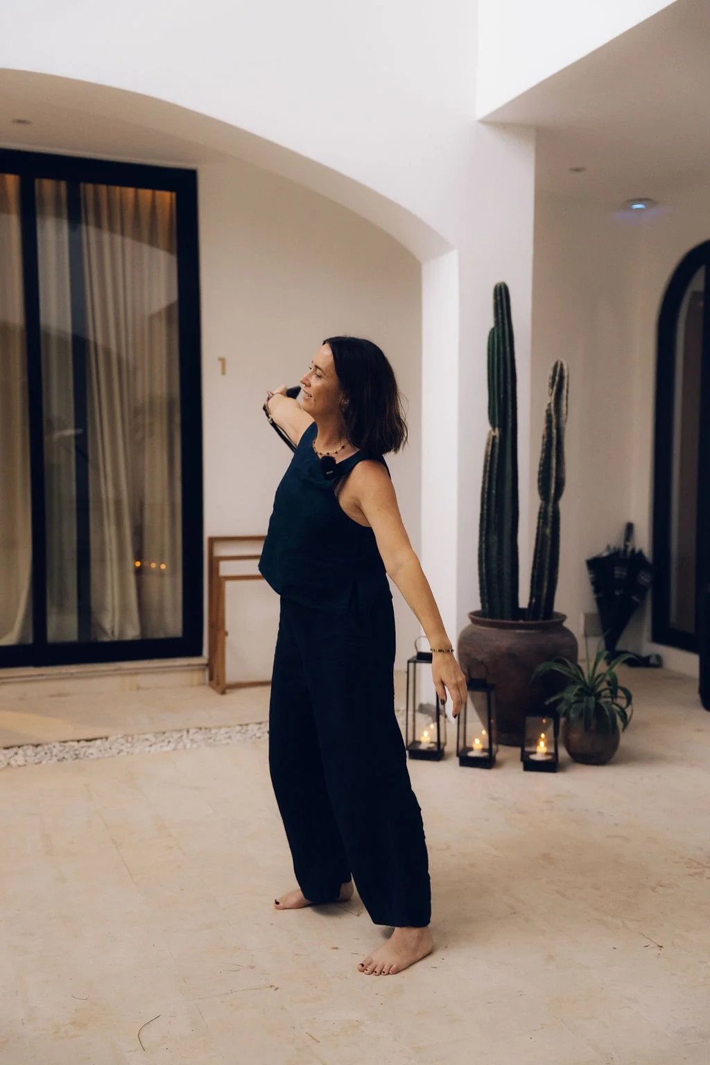 Woman standing barefoot indoors, smiling, with large potted cacti and candles in lanterns behind her.