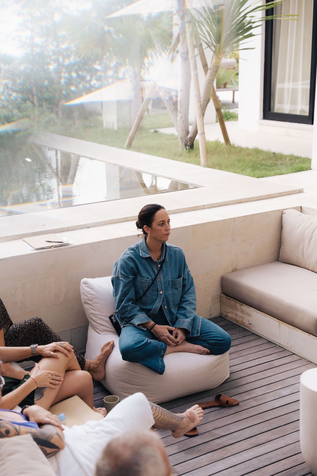 Woman sitting cross-legged on a white cushion indoors, wearing a blue denim jacket and jeans, with a modern outdoor view of trees and grass through large glass windows behind her.