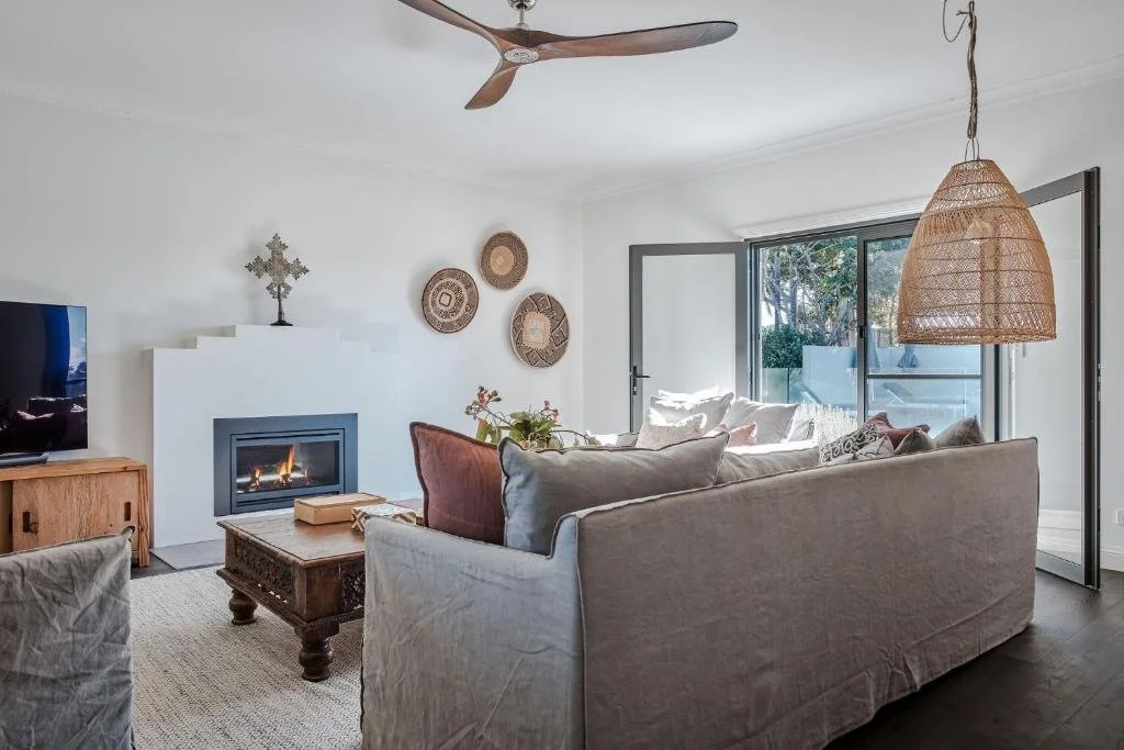 Living room with white walls, a white sofa with pillows, a wooden coffee table, a fireplace, a wooden TV stand with a flat-screen TV, decorative wall hangings, a large window with a door, a woven pendant light, and a ceiling fan.