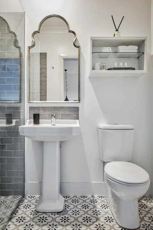 Bathroom with a white pedestal sink, a large mirror, a toilet, and a white wall-mounted shelf holding towels and toiletries.