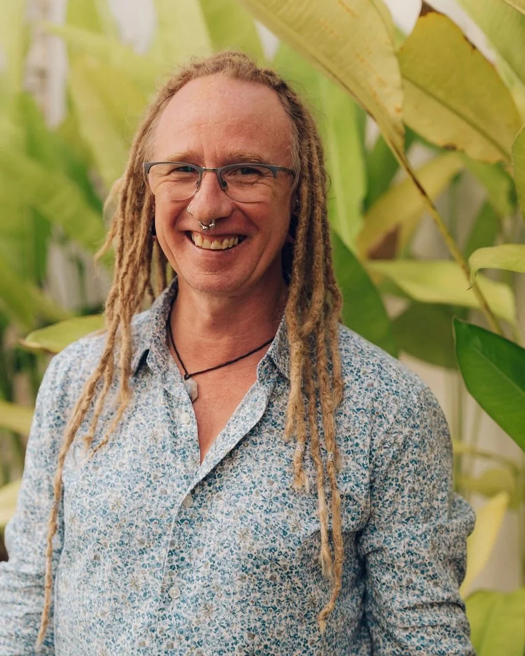A smiling man with light skin, glasses, dreadlocks, and a septum piercing wearing a button-up shirt with a floral pattern, standing among green plants.