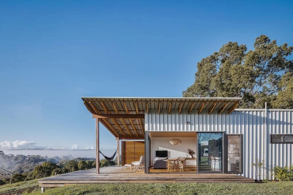 Modern house with an outdoor deck, hammock, and a view of trees and mountains in the background under a clear blue sky.