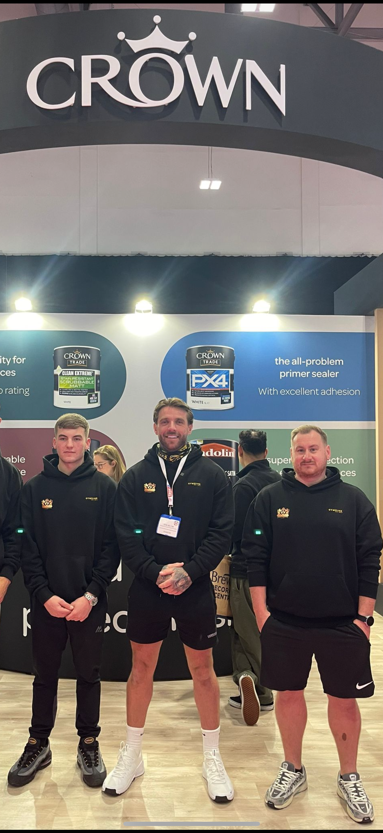 Three men wearing black hoodies with a crown logo, standing in front of a display at a trade show. The display features Crown trade paint cans and promotional text.