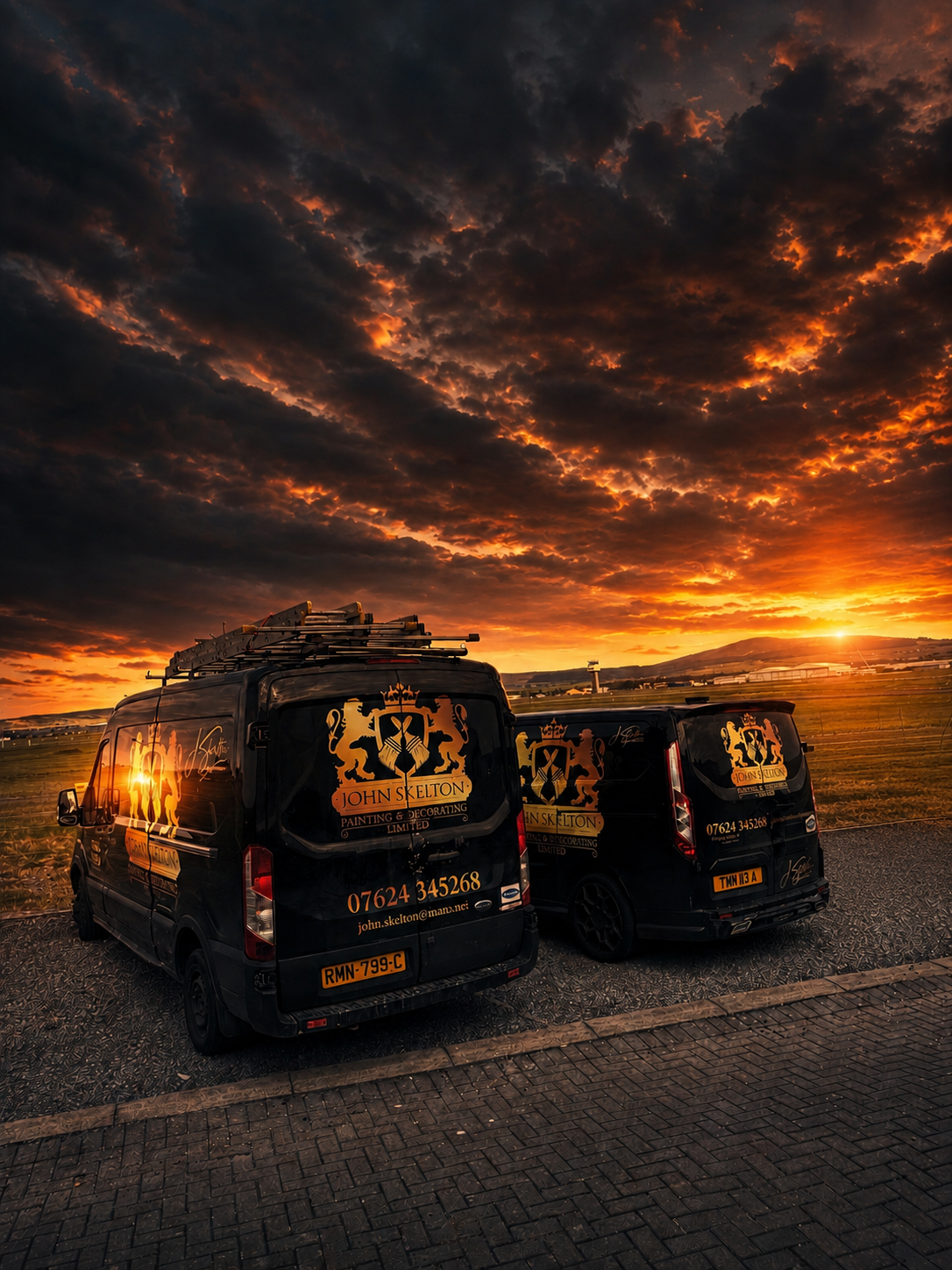 Two black vans parked on a paved surface with a scenic sunset sky and clouds in the background. The vans display the logo and contact information for John Skelton Painting & Decorating.