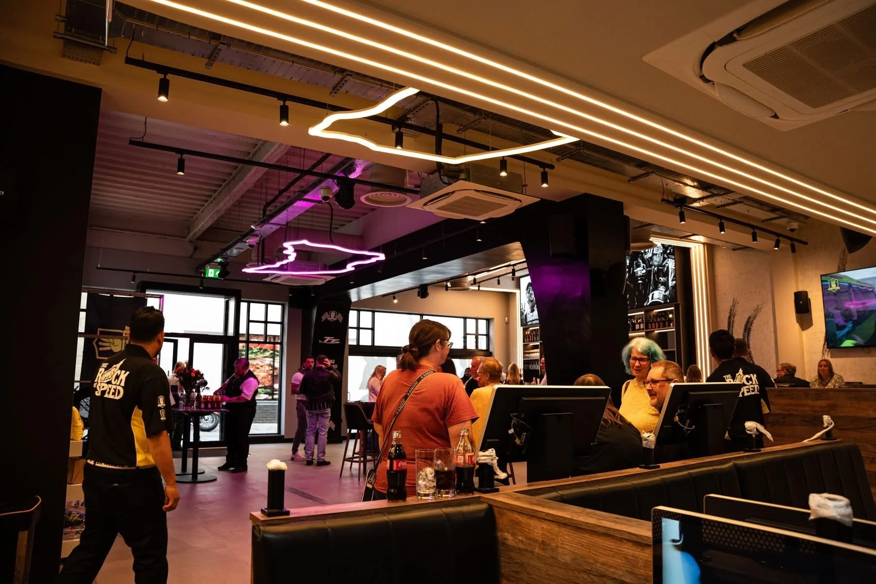 People gather in a modern restaurant with neon light decorations on the ceiling. Some are sitting and talking, others are standing near the bar area. The interior features black and wood accents, with large windows and a television screen on the wall