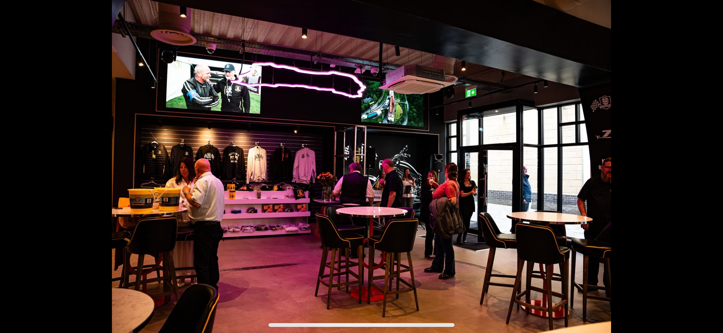 Interior of a lively retail store or cafe with people chatting and browsing. Display shelves with folded clothes and jackets, neon signage, and large windows letting in natural light.