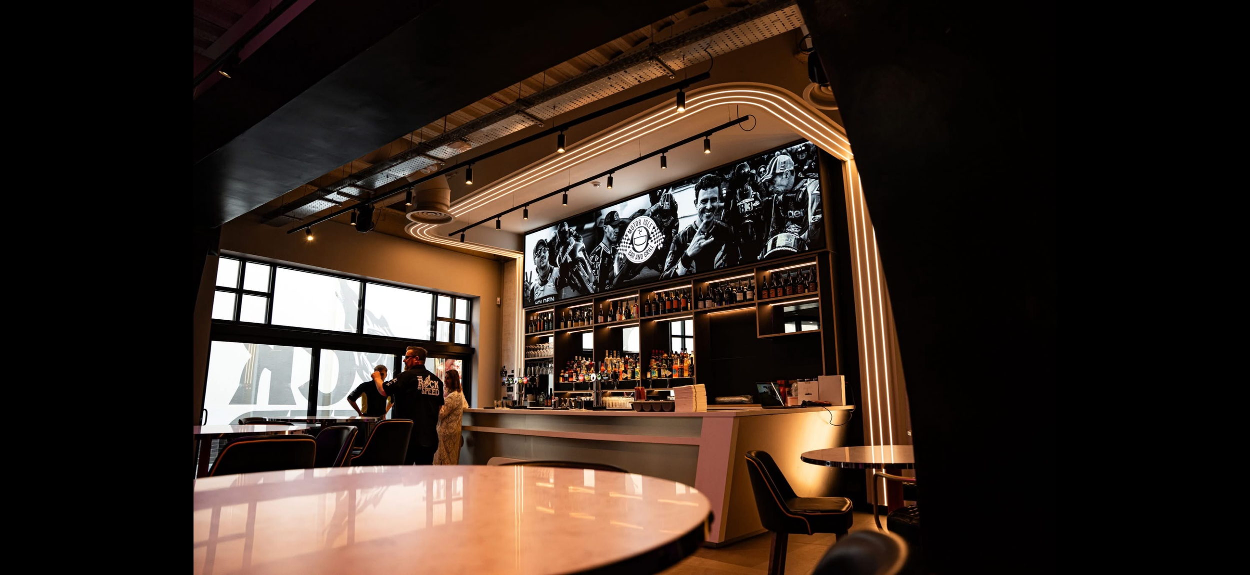 Interior of a modern bar and restaurant with a large black-and-white photo of motorcyclists above the bar, bottles of alcohol on shelves, and seating at tables and a bar counter.