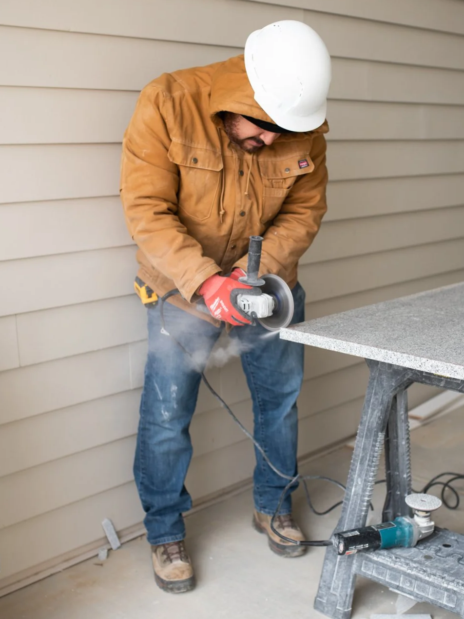 A man wearing a yellow jacket, hard hat, sunglasses, and gloves uses a power angle grinder on a countertop, with dust and sparks visible, in an outdoor workspace with beige siding.