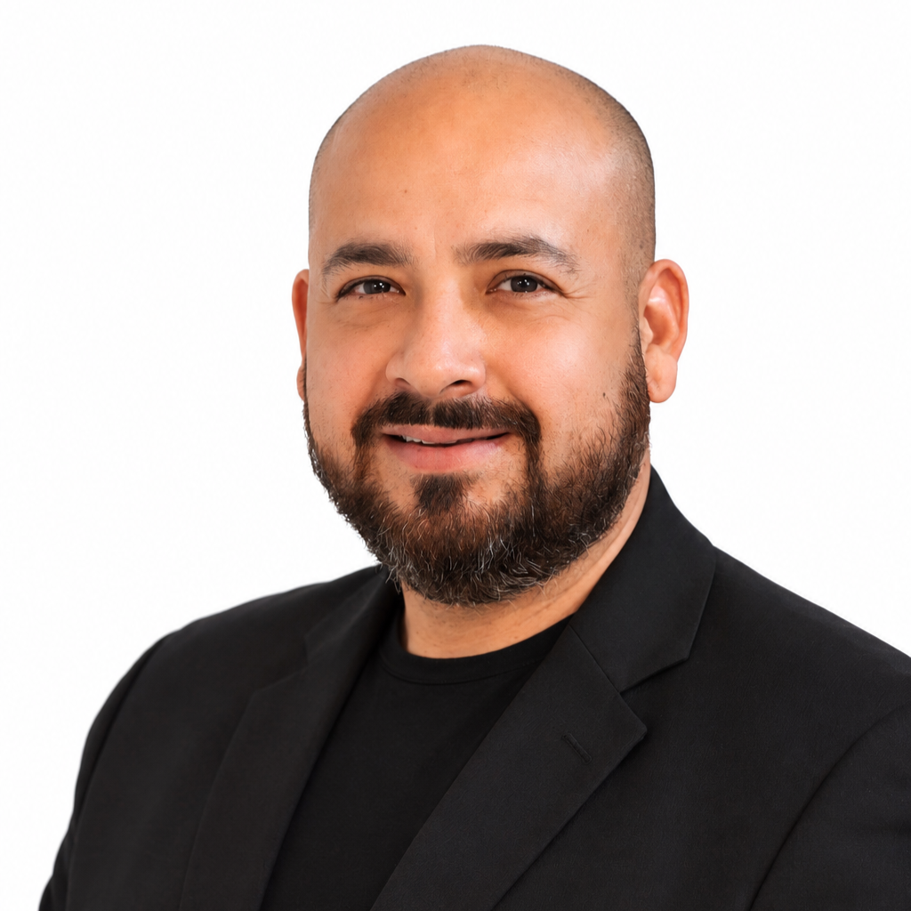 A professional man with a bald head, beard, and mustache, wearing a black blazer over a black shirt, smiling against a white background.