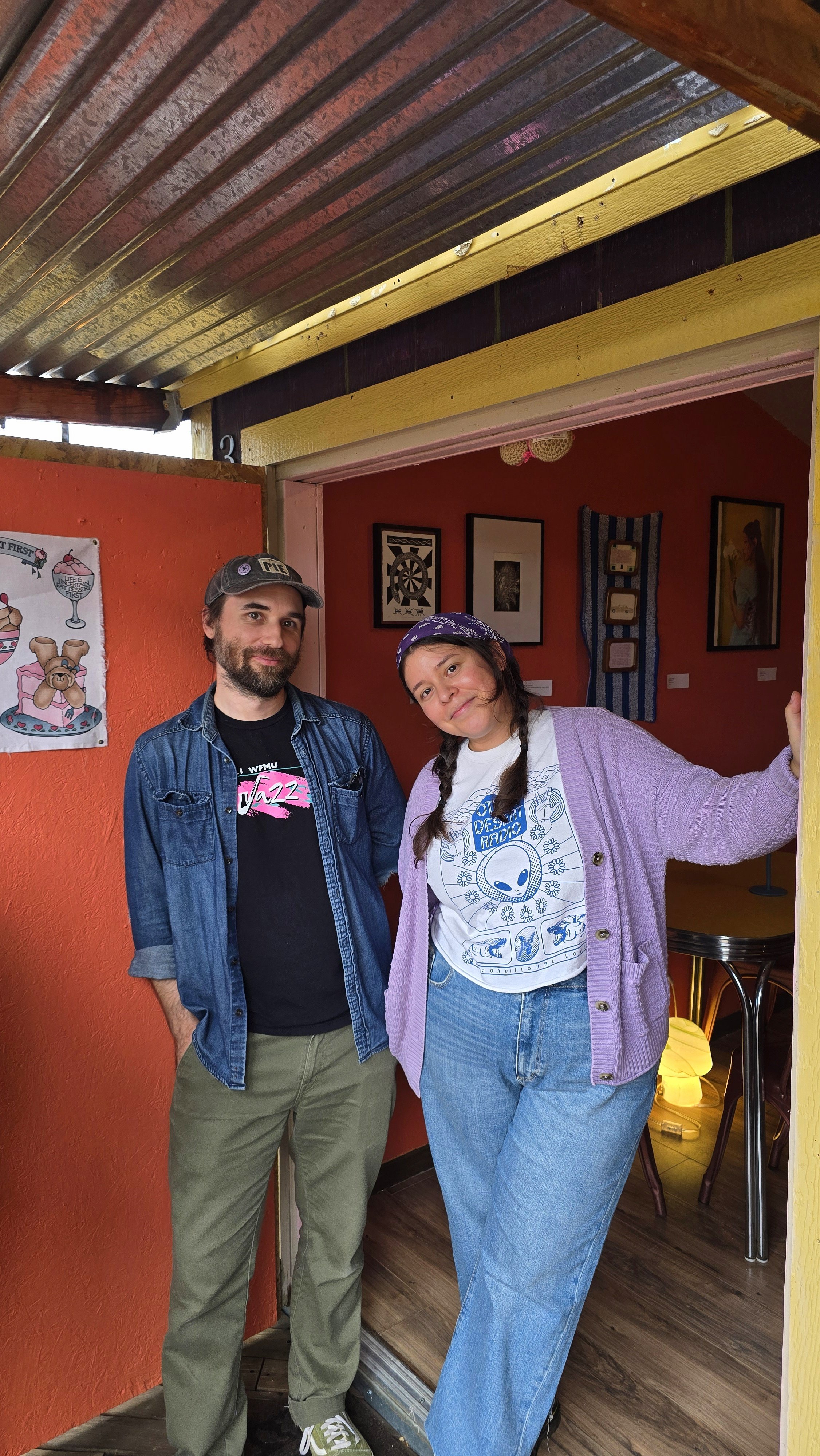 Two people standing in a doorway inside an art gallery, with framed artwork on the walls behind them. One man has a beard, wearing a baseball cap, a denim jacket, and cargo pants. The woman has long hair in braids, wearing a purple cardigan, a graphic t-shirt, and jeans. They are smiling at the camera.