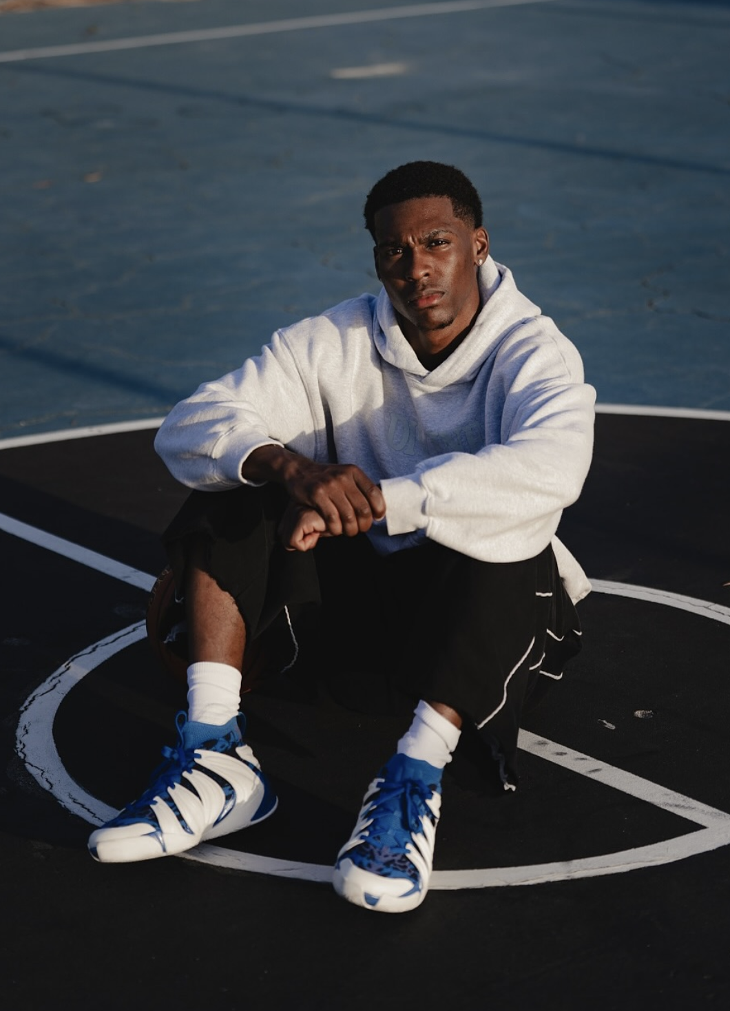 Young man with blonde hair sitting on a black stool, wearing a colorful striped Polo shirt, denim shorts, white sneakers, and white socks, holding a basketball against a solid mustard yellow background.