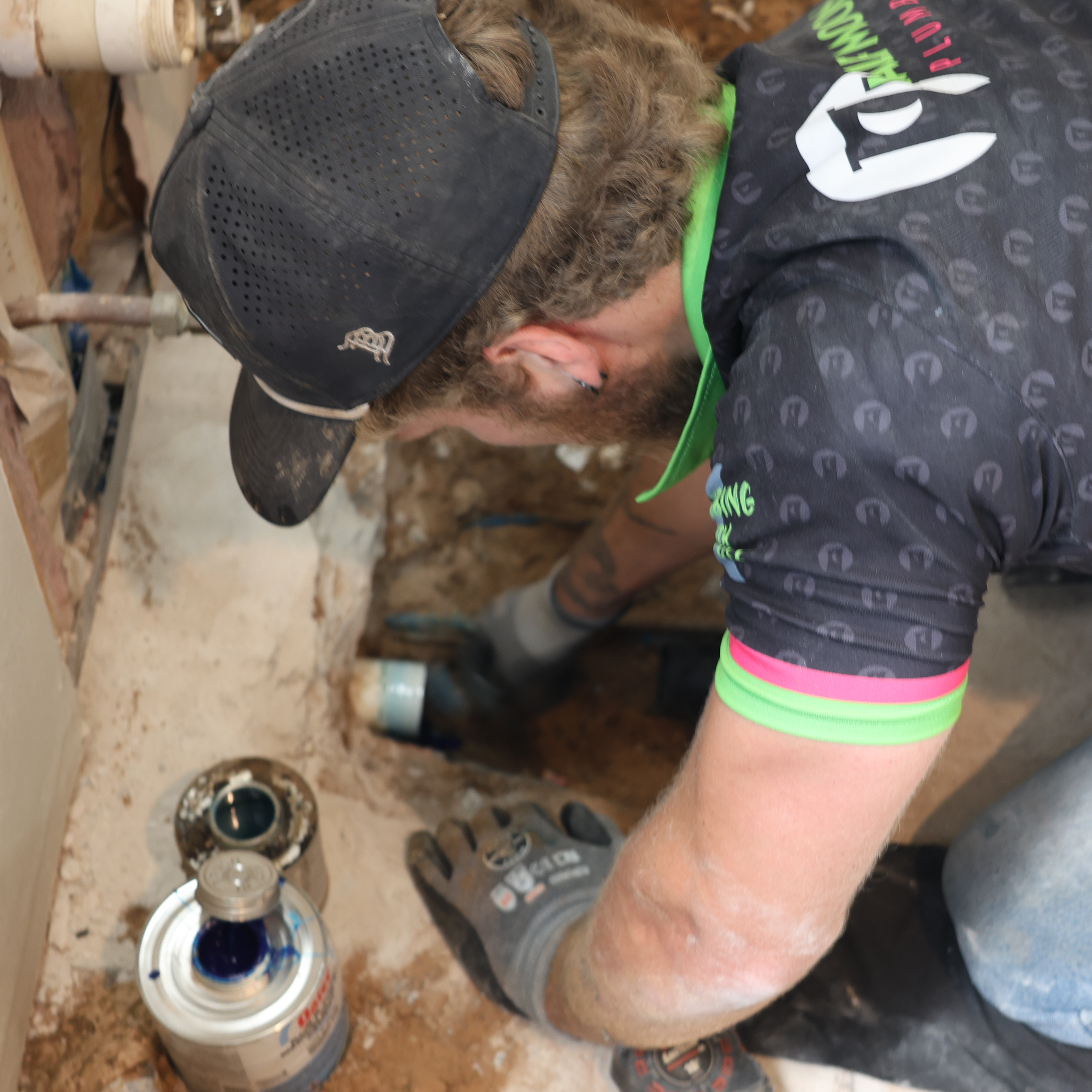 Person doing plumbing work in a small space, wearing gloves and a cap, with plumbing tools and materials nearby.