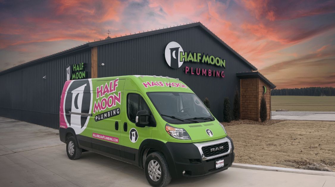 A bright green plumbing service van parked outside a building with a large sign that reads "Half Moon Plumbing." The van and building logos feature a half moon symbol.