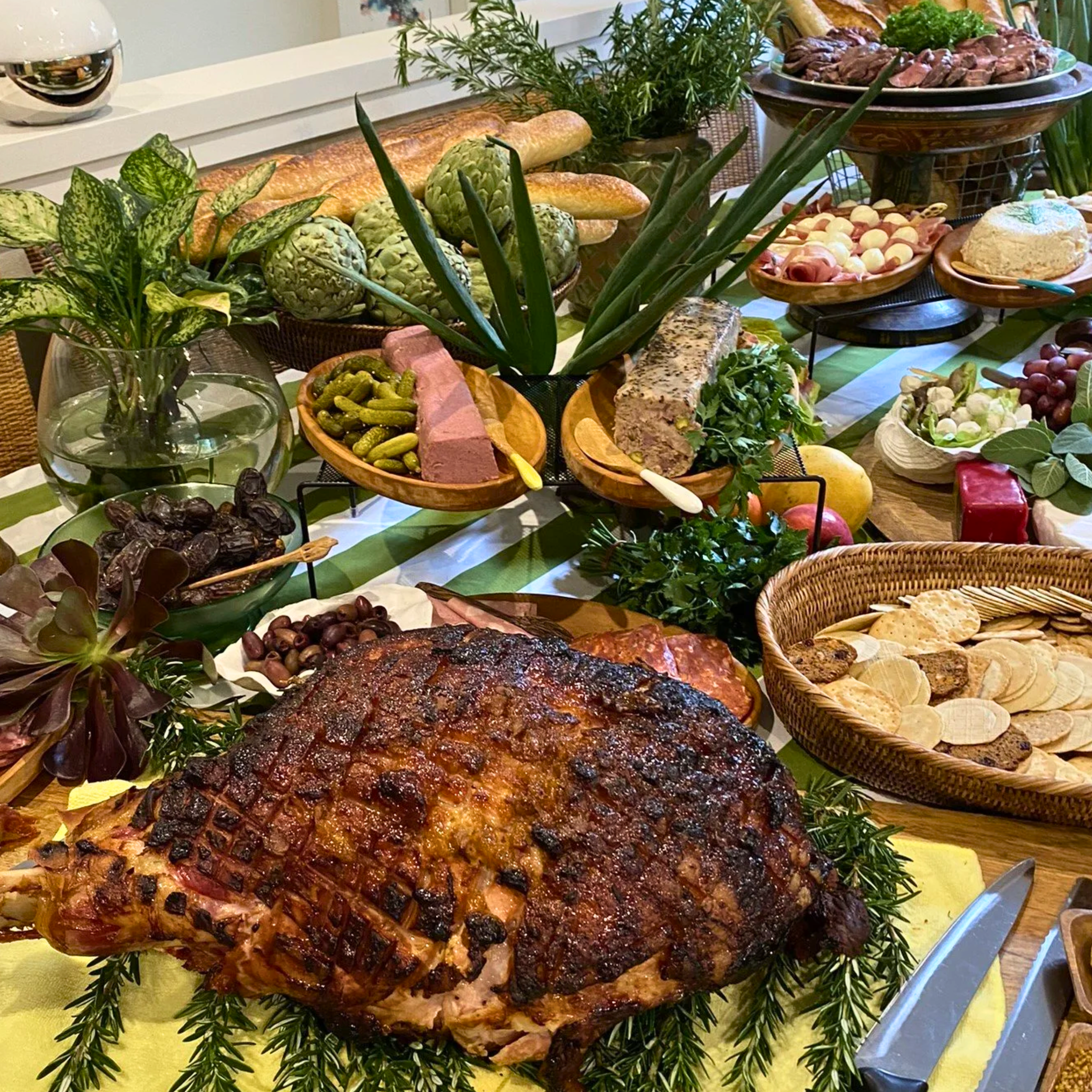 A table decorated with various foods including a glazed ham, cheese, crackers, grapes, pickles, bread, and vegetables, along with greenery and flowers for decoration.